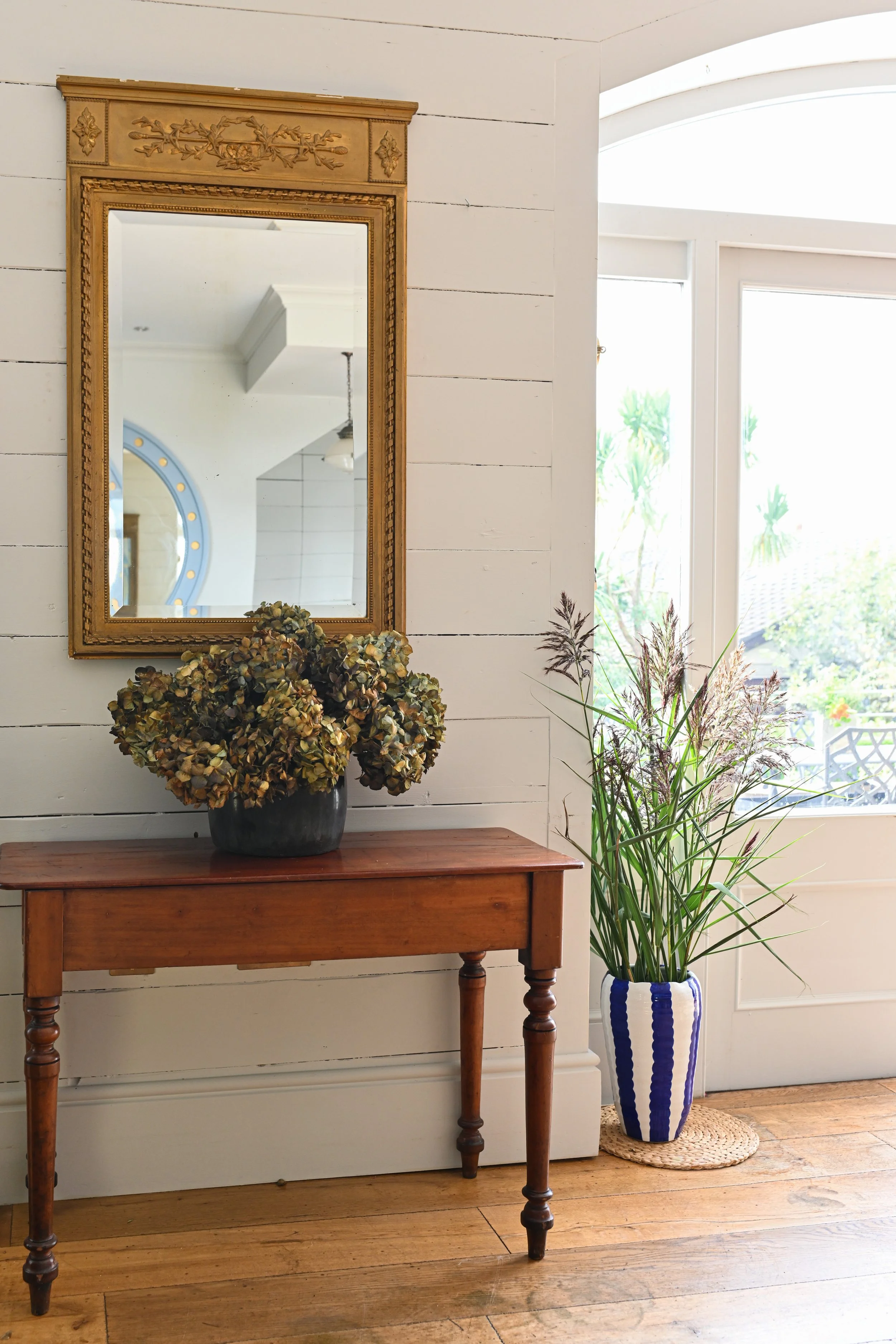 A wooden console table with a potted dried hydrangea on top, a large mirror with a gold frame hung above, and a tall vase with grass and other plants next to a window showing a sunny outdoor scene.