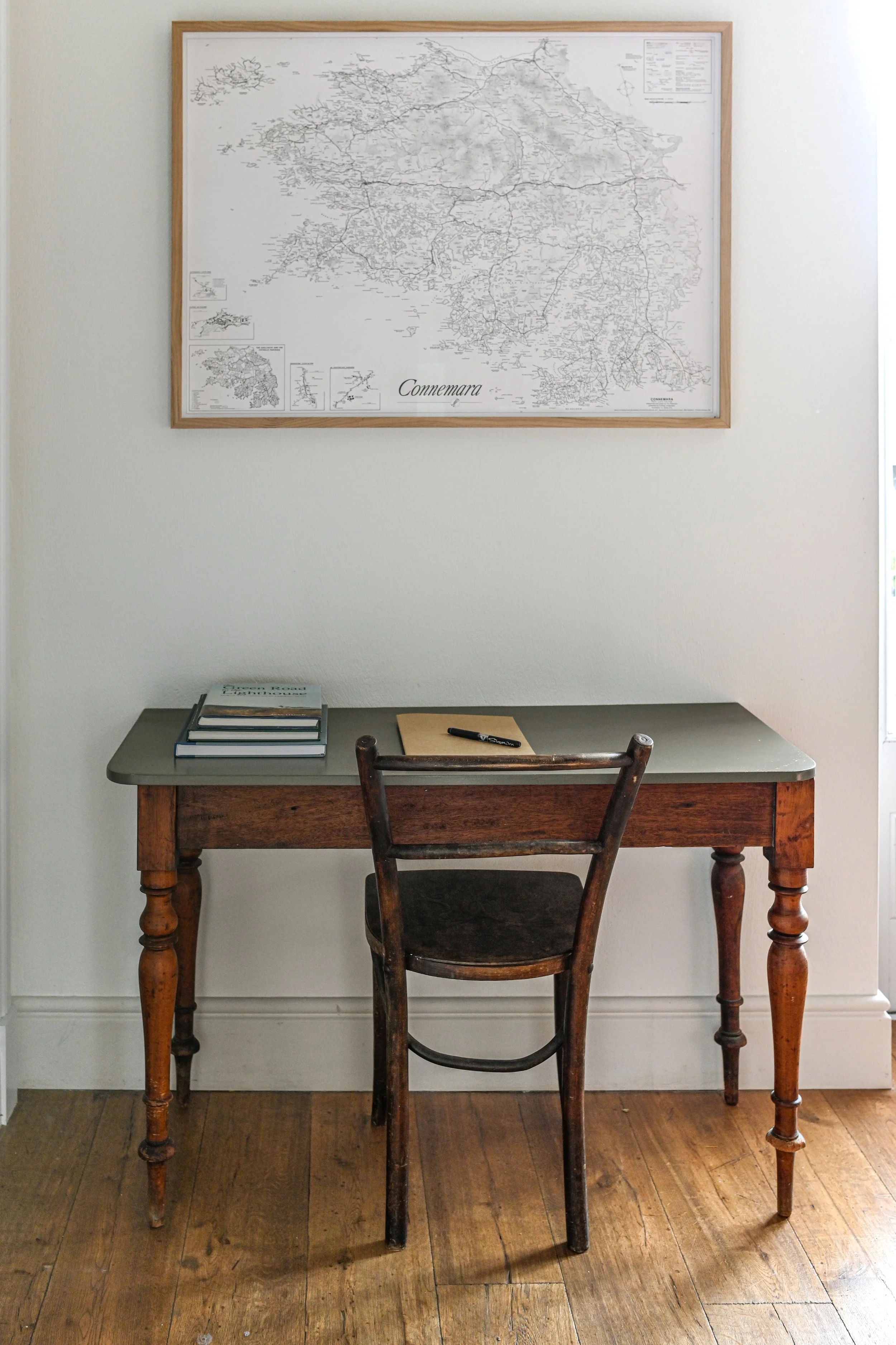 A vintage wooden desk with a gray top surface, a chair, a folded notebook, and a black pen on top, positioned against a white wall with a framed map hanging above the desk.