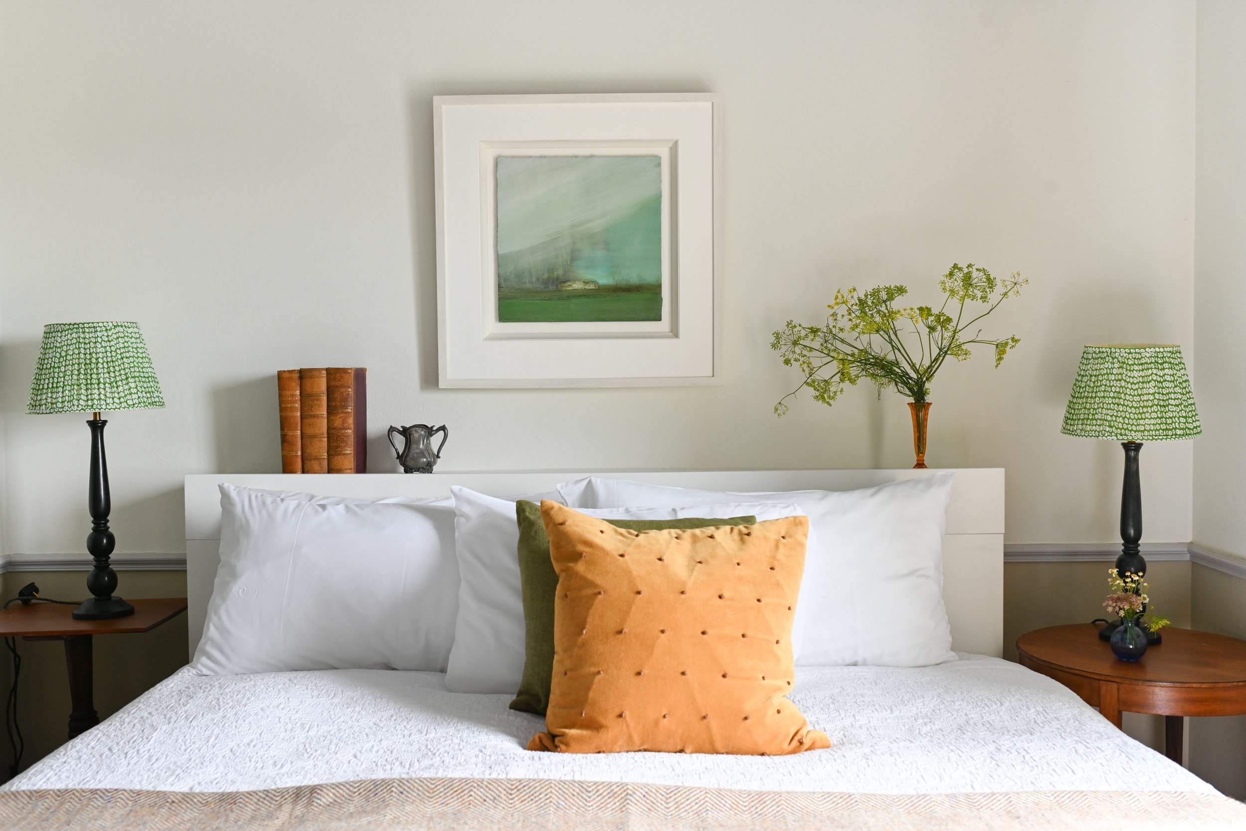 A neatly made bed with white linens, decorative pillows, two green lamps on bedside tables, and wall art with greenery and sky in the background.