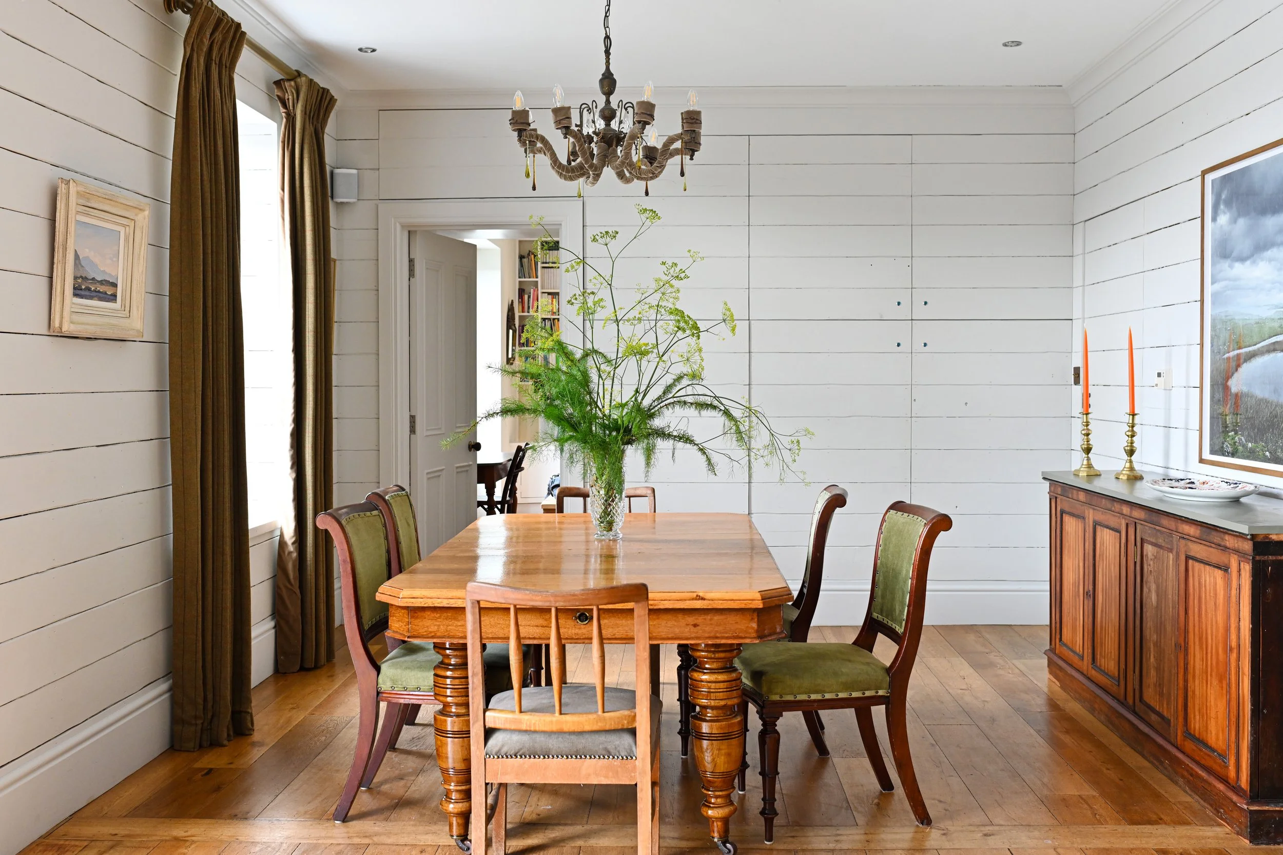 Dining room with a wooden table, six chairs, a large vase with green plants, a chandelier, a wooden sideboard with two orange candlesticks, framed landscape paintings, and curtains next to a window.
