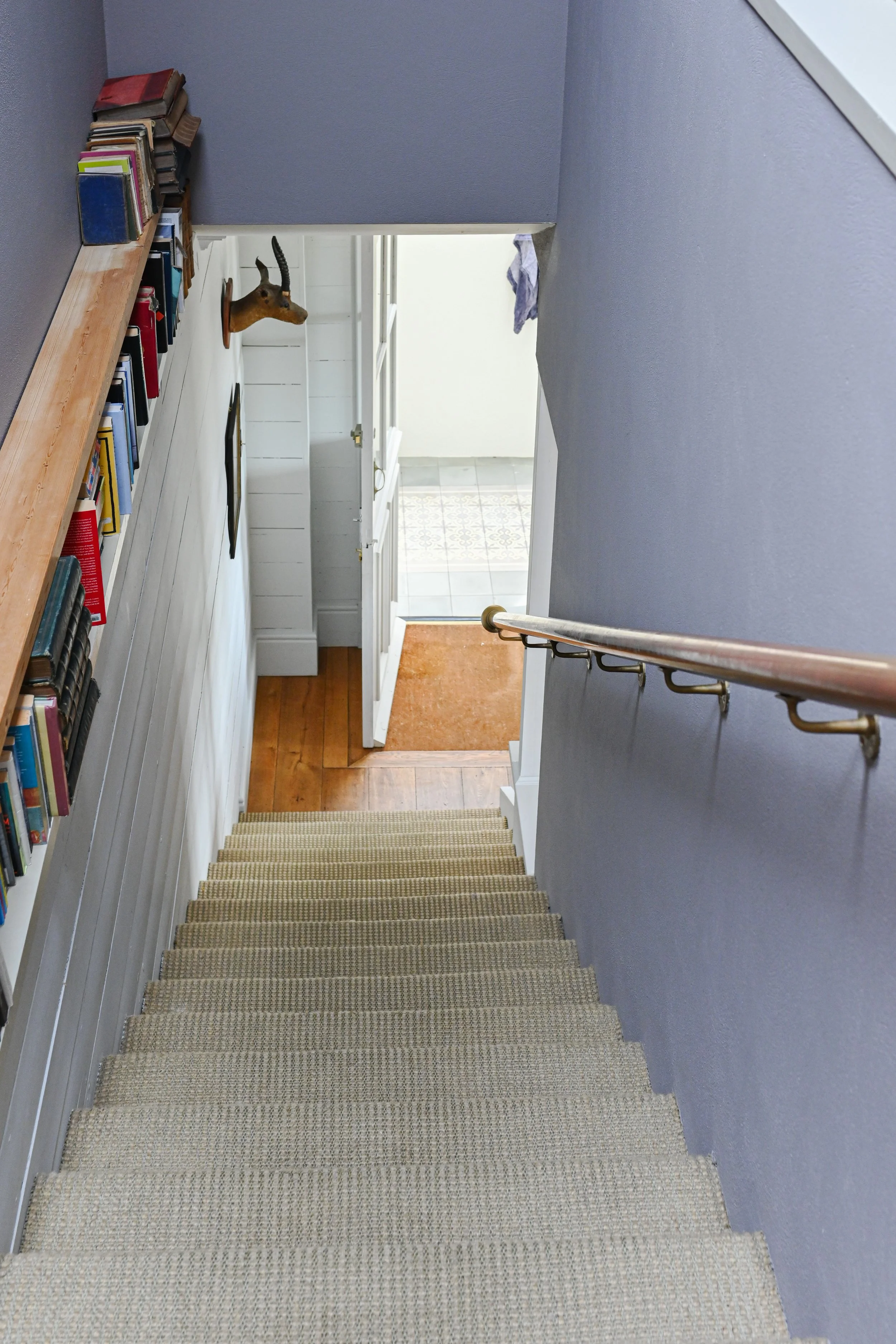 A staircase leading down to a small landing with an open door to a tile-floored room. A bookshelf filled with books is visible on the left wall, and a mounted animal head is on the wall near the doorway.