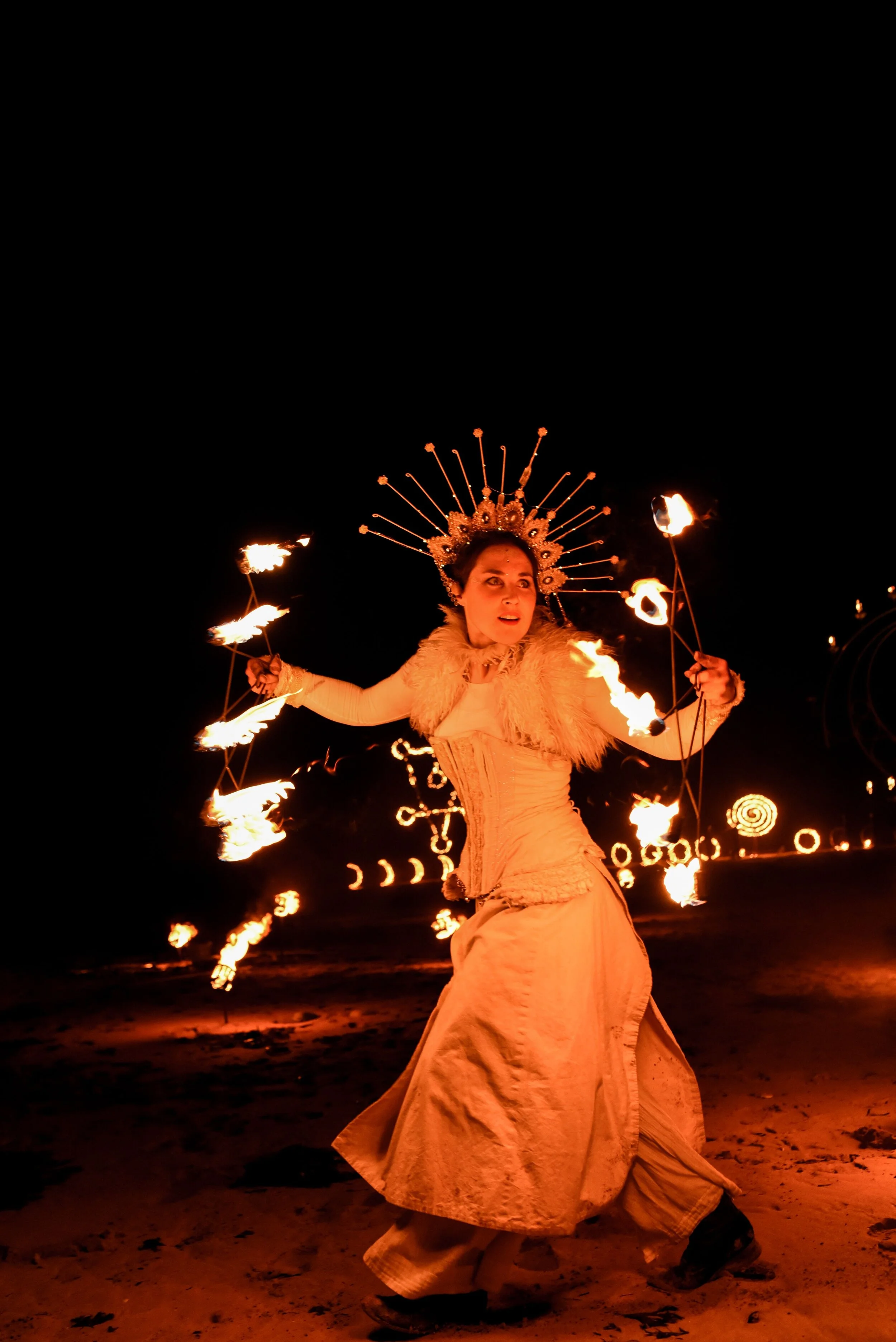 Woman performing fire dance at night, dressed in white costume with a decorative headdress, spinning fire sticks against a dark background.