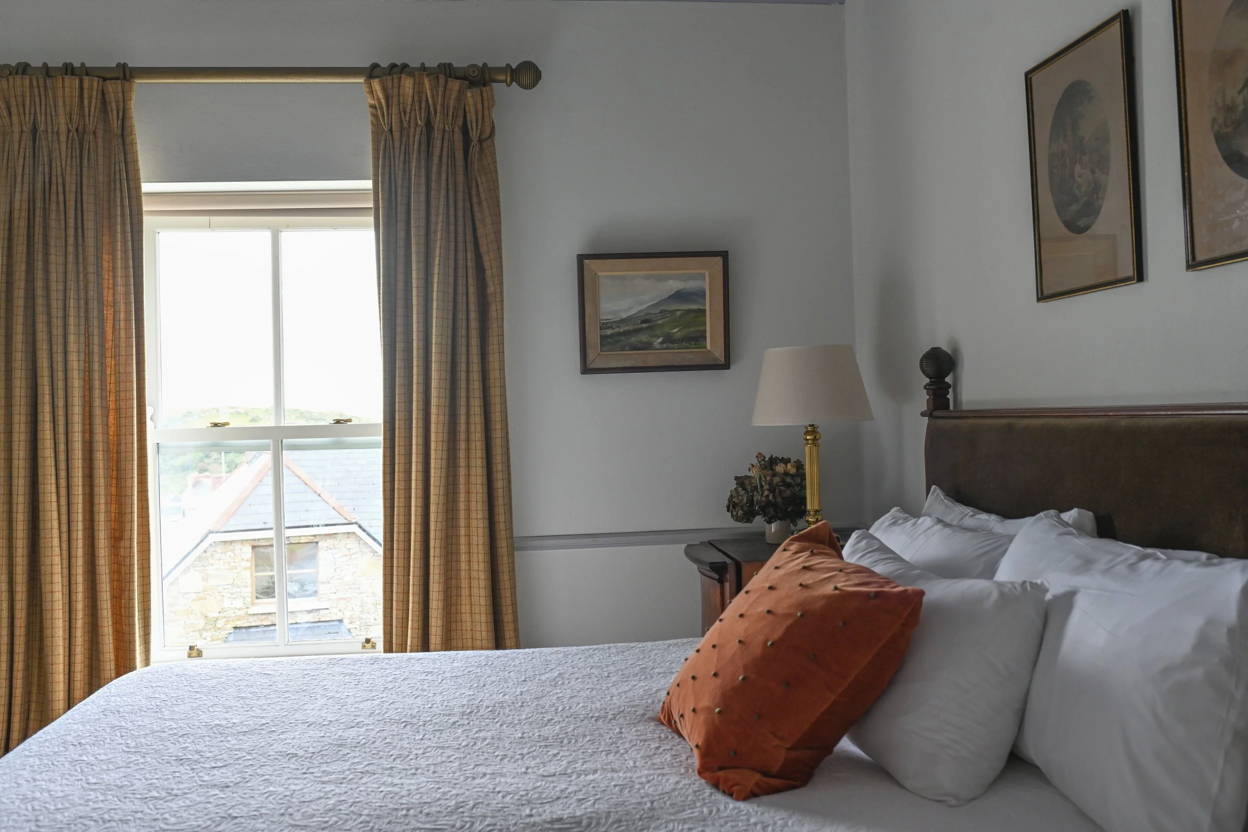 A cozy bedroom with a large window with plaid curtains, a bed with white bedding and an orange pillow, a wooden nightstand with a lamp and decorative flowers, and framed landscape paintings on the white walls.