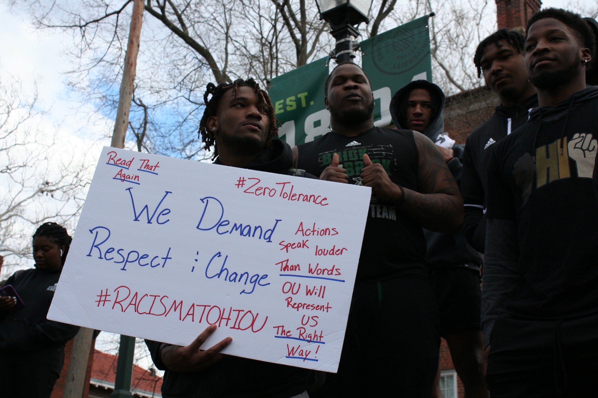  A group of Ohio University football players speak and stand in solidarity with protesters. A member of the team is holding a sign that reads, “Read That Again: We Demand Respect and Change #RACISMATOHIOU” along with “#ZeroTolerance,” “Actions speak 