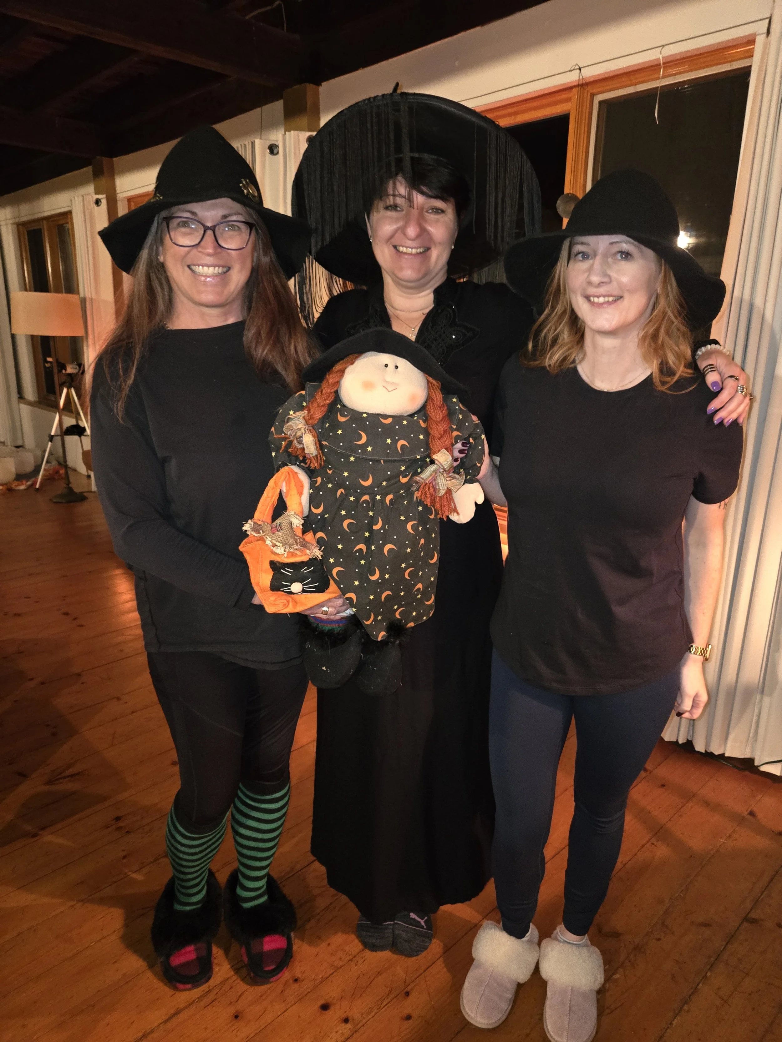 Three women wearing witch hats holding a doll witch smiling.