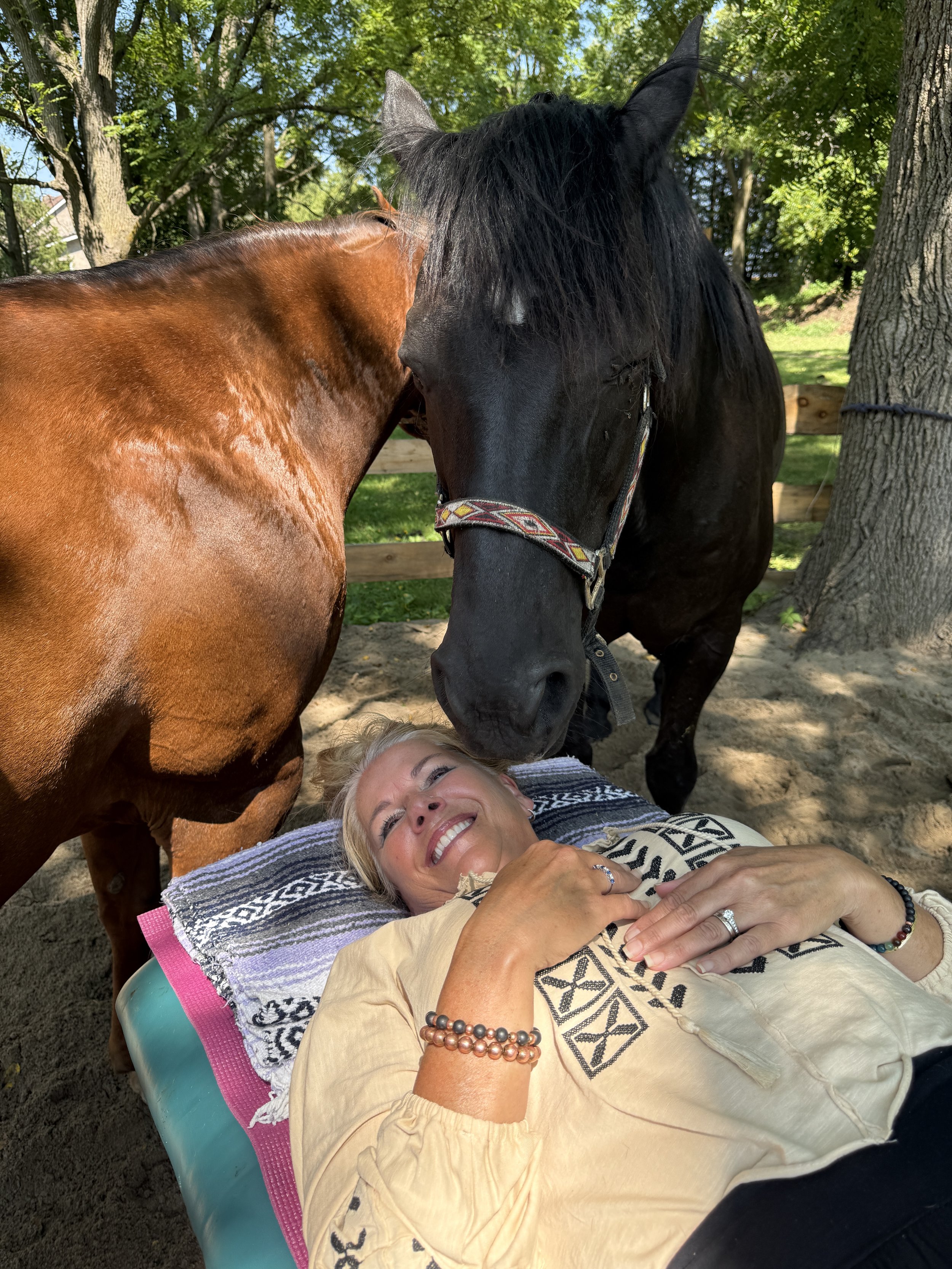 Woman laying on a table smiling as two horses stand near her head. One is touching her with her nose.
