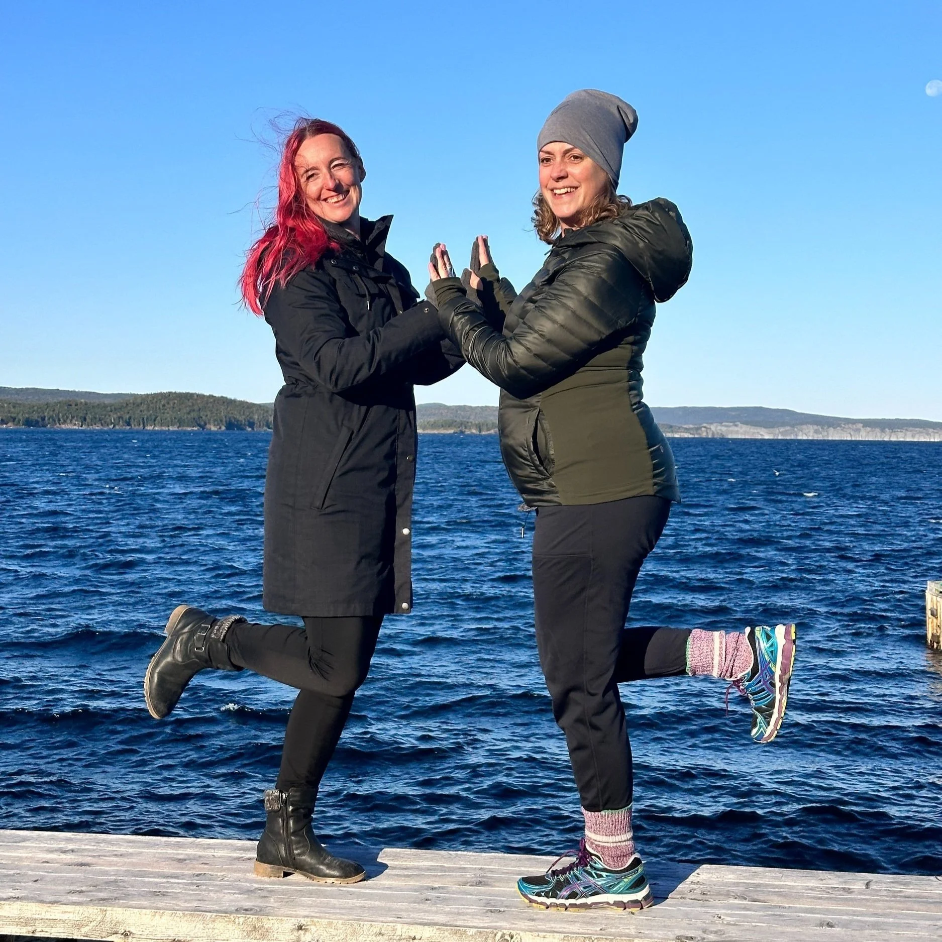 Retreat hosts Catherine & Jessica standing on a table facing each other with the blue ocean behind them.