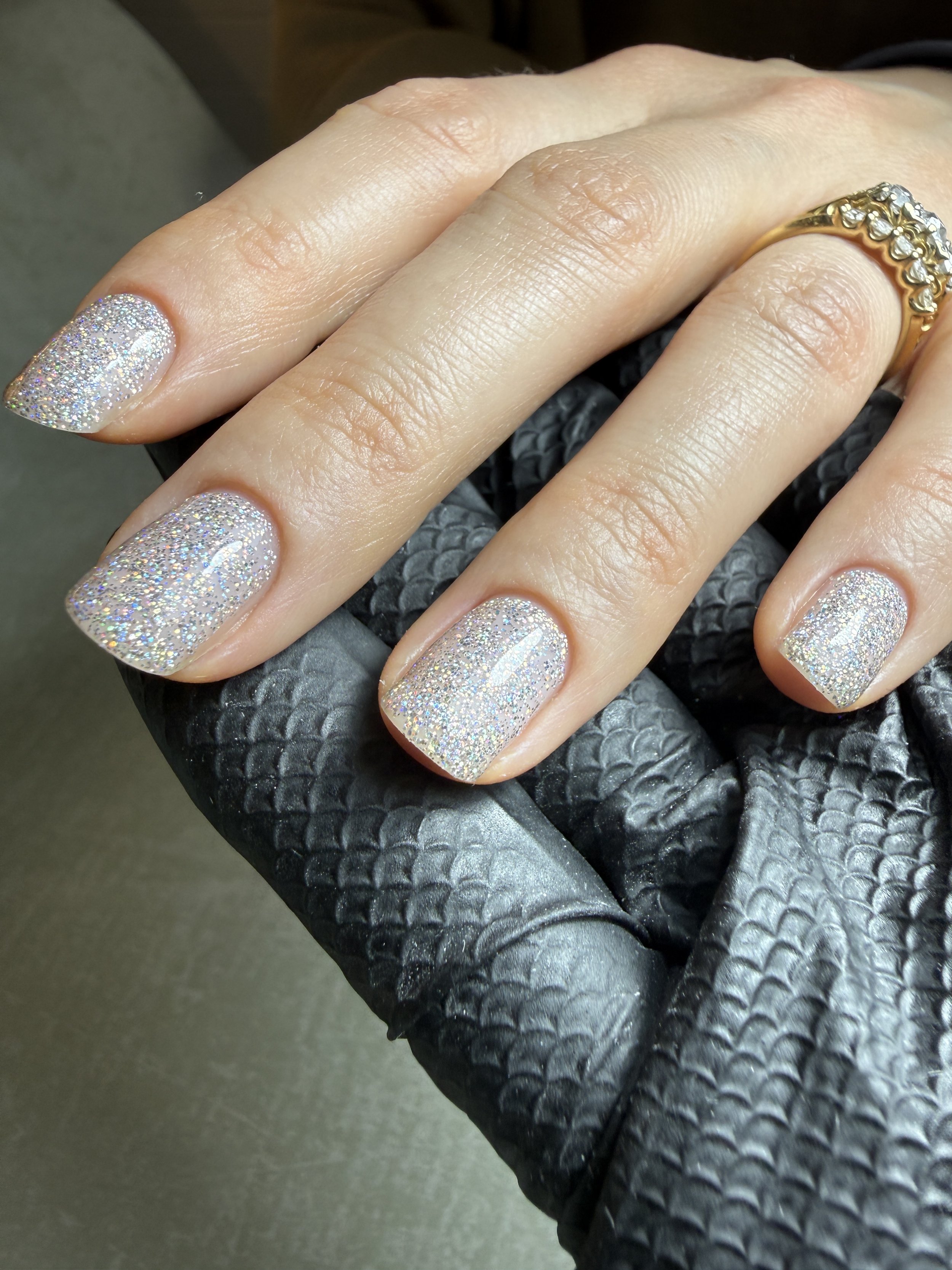 Close-up of a hand with glittery nail polish on fingernails, gold ring with diamonds on ring finger, holding a black textured leather surface.