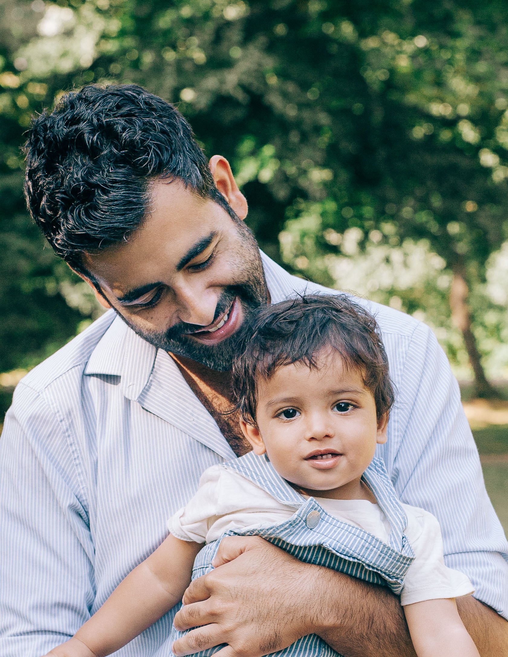 South Asian Dad looking down hugging his toddler son in the park