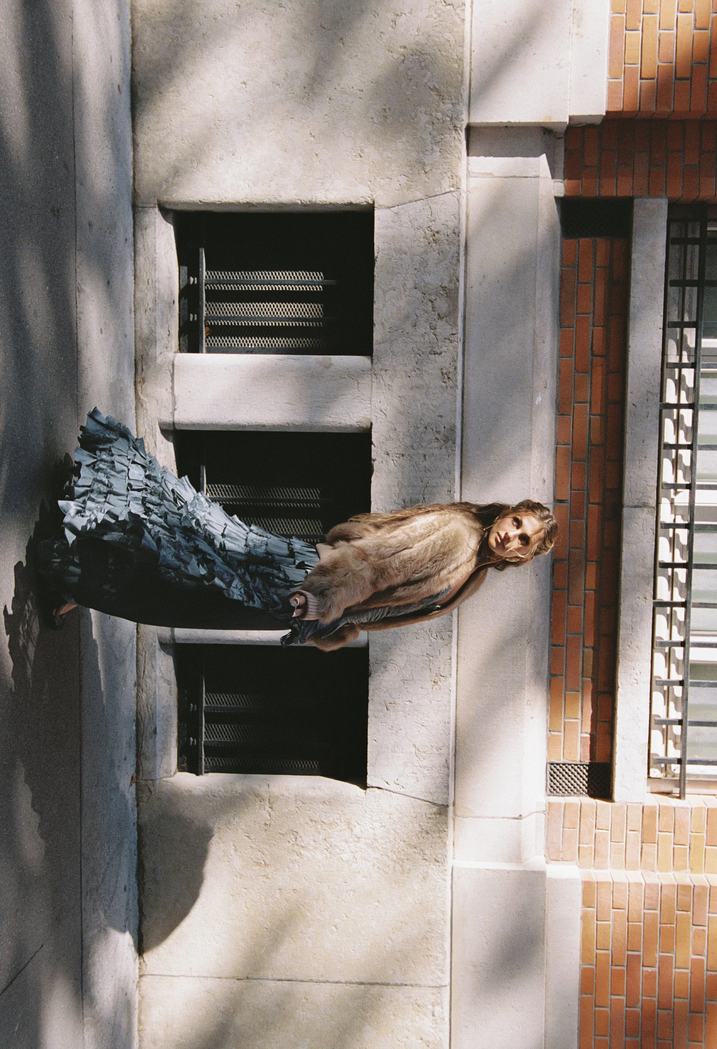 A woman in a fur coat and patterned pants standing outdoors against a concrete wall and brick structure, with sunlight and shadows.