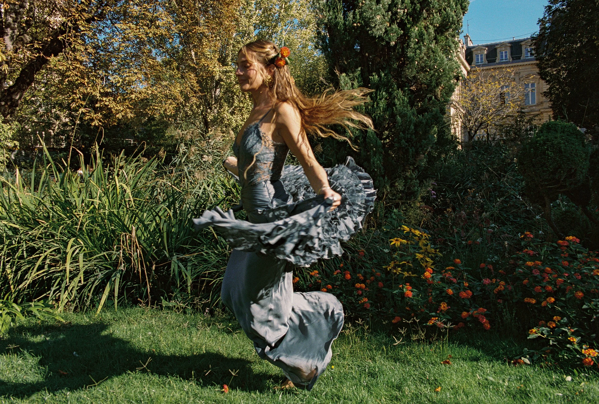 A woman with long hair, decorated with orange flowers, stands outdoors among trees and plants, wearing a gray dress, with a large gray fabric or bag around her waist, and appears to be enjoying a windy day.
