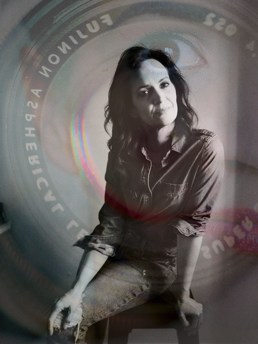 A black-and-white photo of a woman with dark wavy hair wearing a denim shirt, sitting on a chair with one leg crossed over the other. There is a reflection of a camera lens overlaying the image.