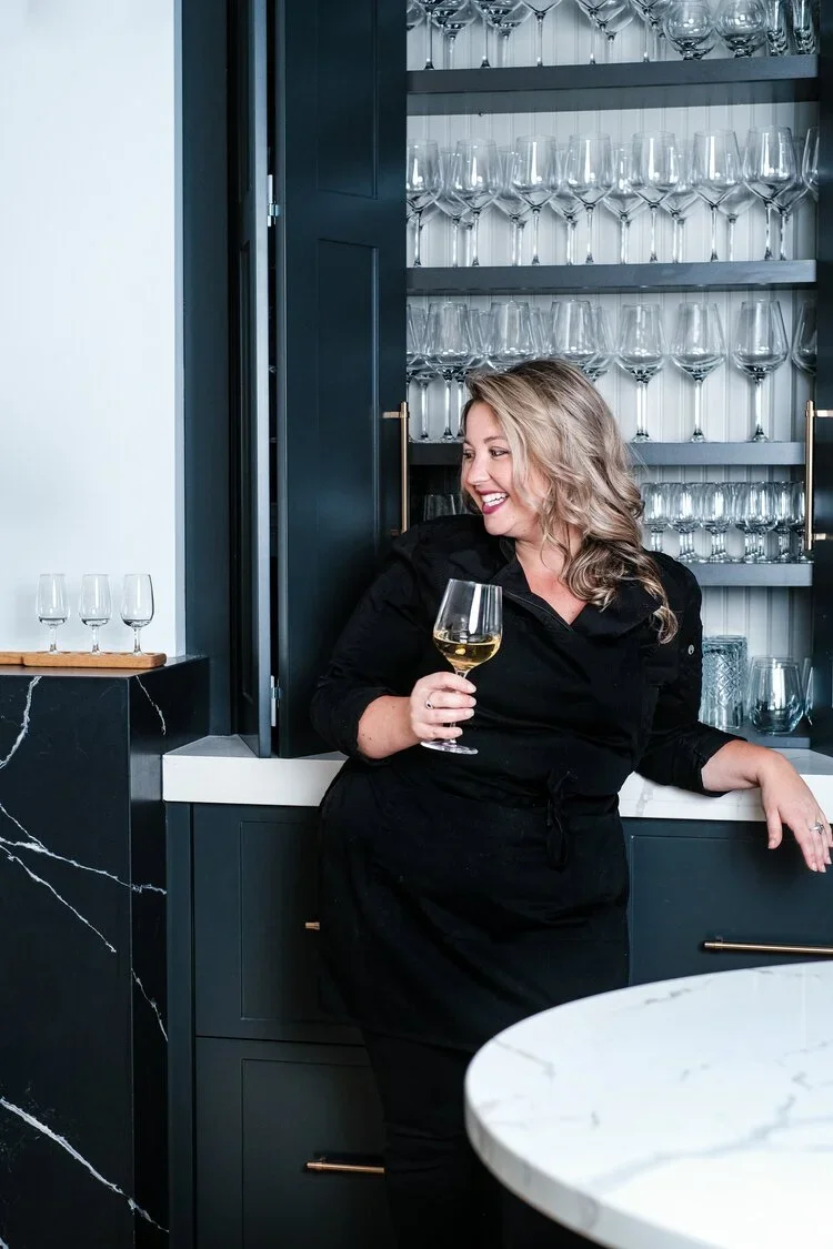 woman in chefs clothing holding wine in front of a cupboard of wine glasses