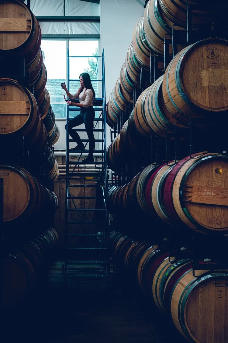 woman climbing a large ladder in a winery pouring wine from a barrel into a glass