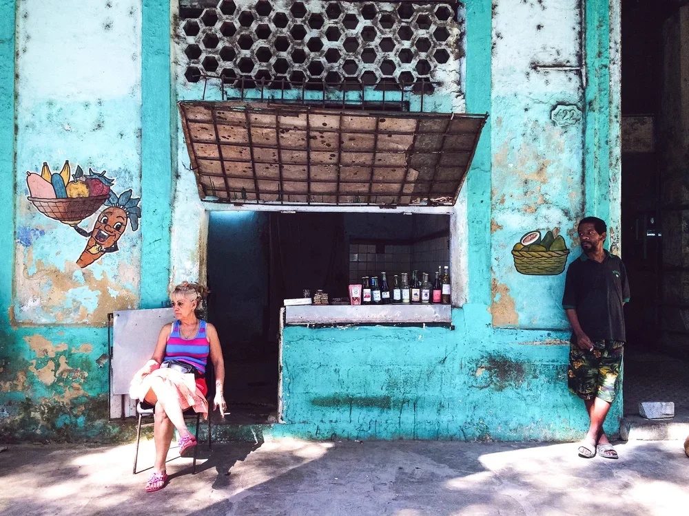 woman smoking a cigarette sitting in a chair outside a fruit market in Havana, Cuba while a man looks at her.