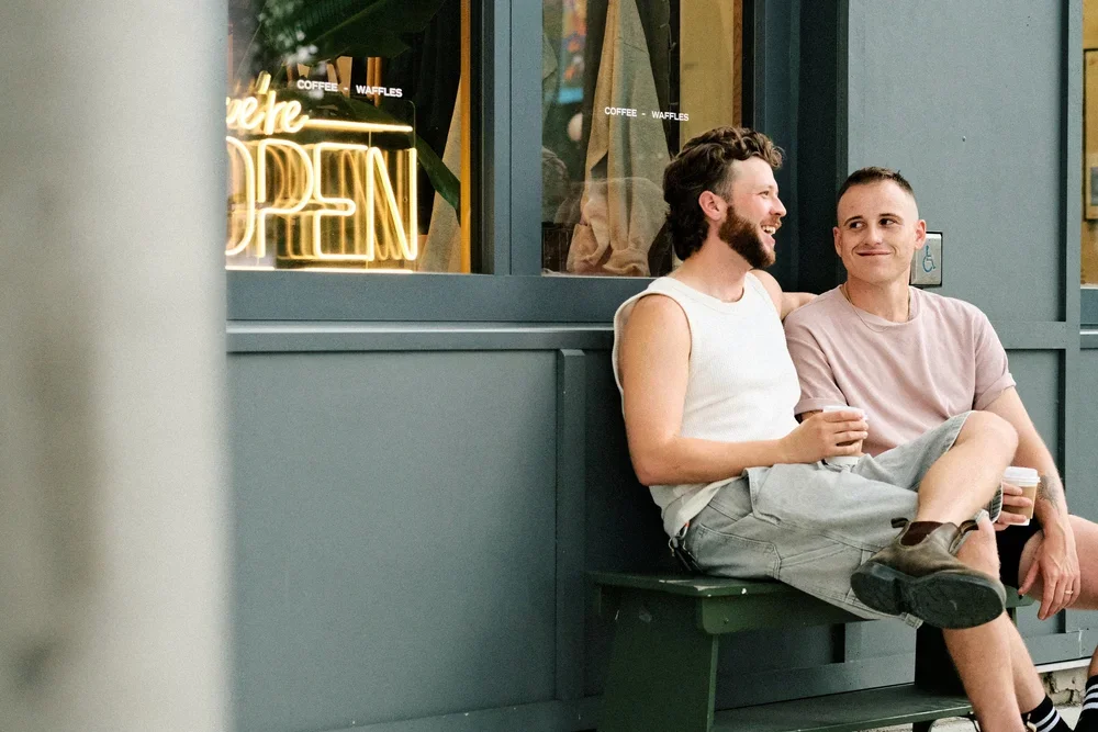 two men sitting outside a cafe on a bench smiling and drinking coffee