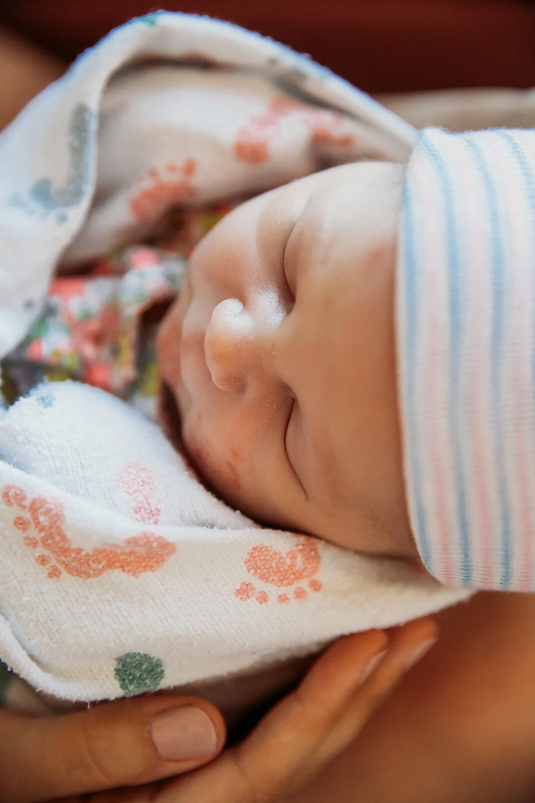 close up of newborn baby's face from above, baby is wrapped in a footprint blanket and pink and blue striped hat issued by hospital