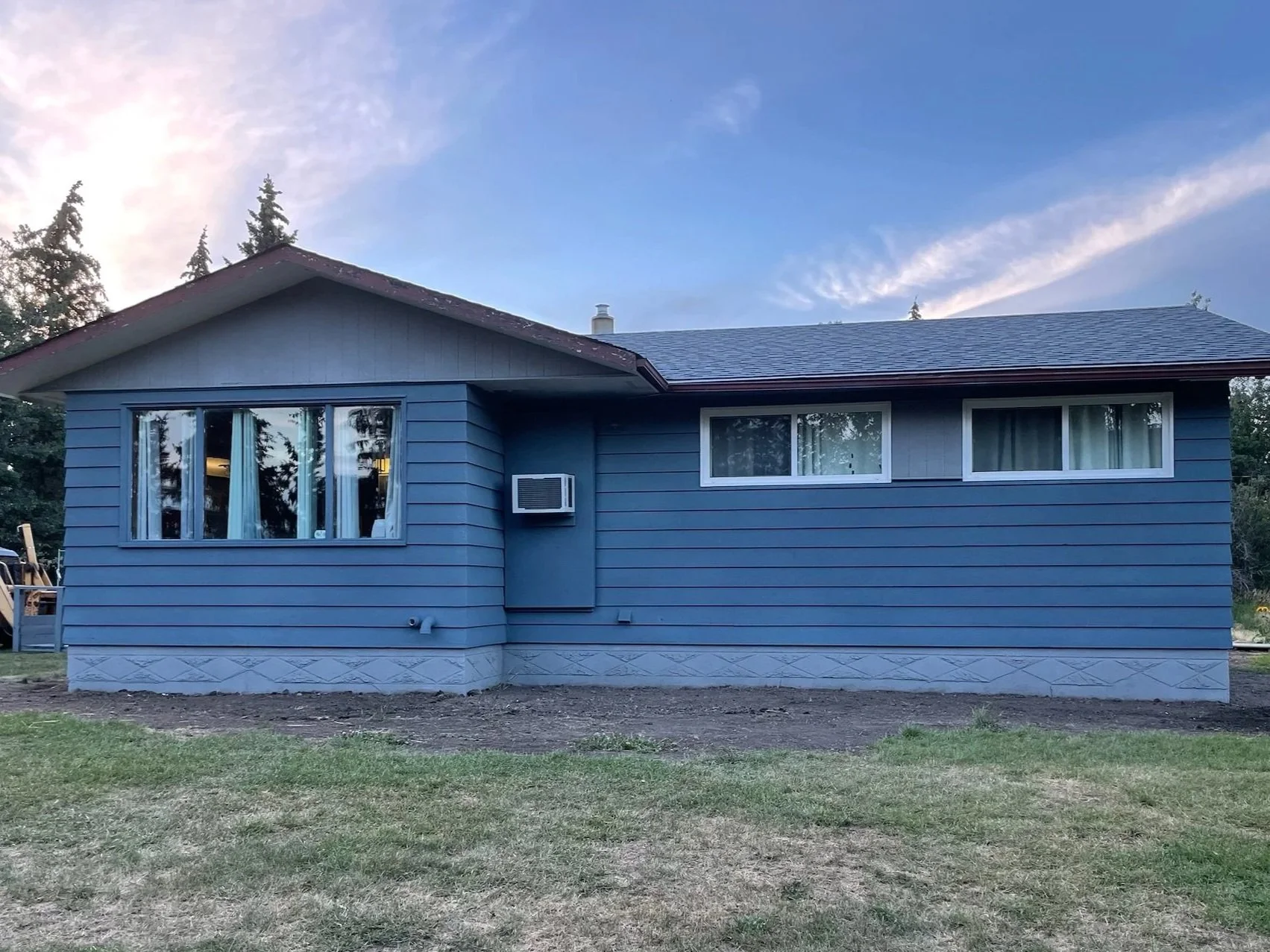 A two-story house with horizontal siding, three windows, an air conditioning unit, and a small chimney, surrounded by a grassy yard and trees, under a partly cloudy, sunset sky. Renovated metal siding with CorkShield cork spray by Vipeq.