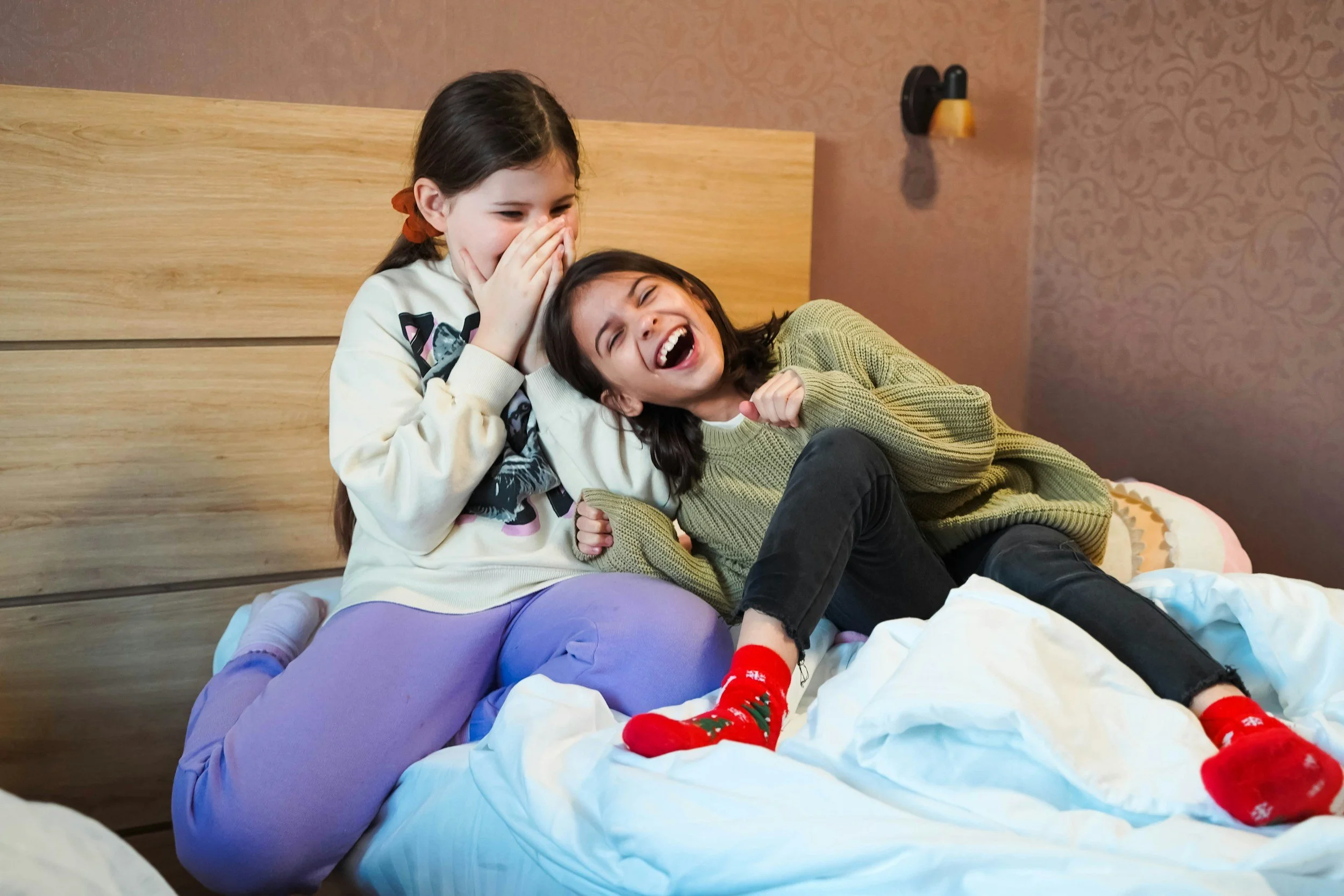 two young girls sit on a bed, laughing together.