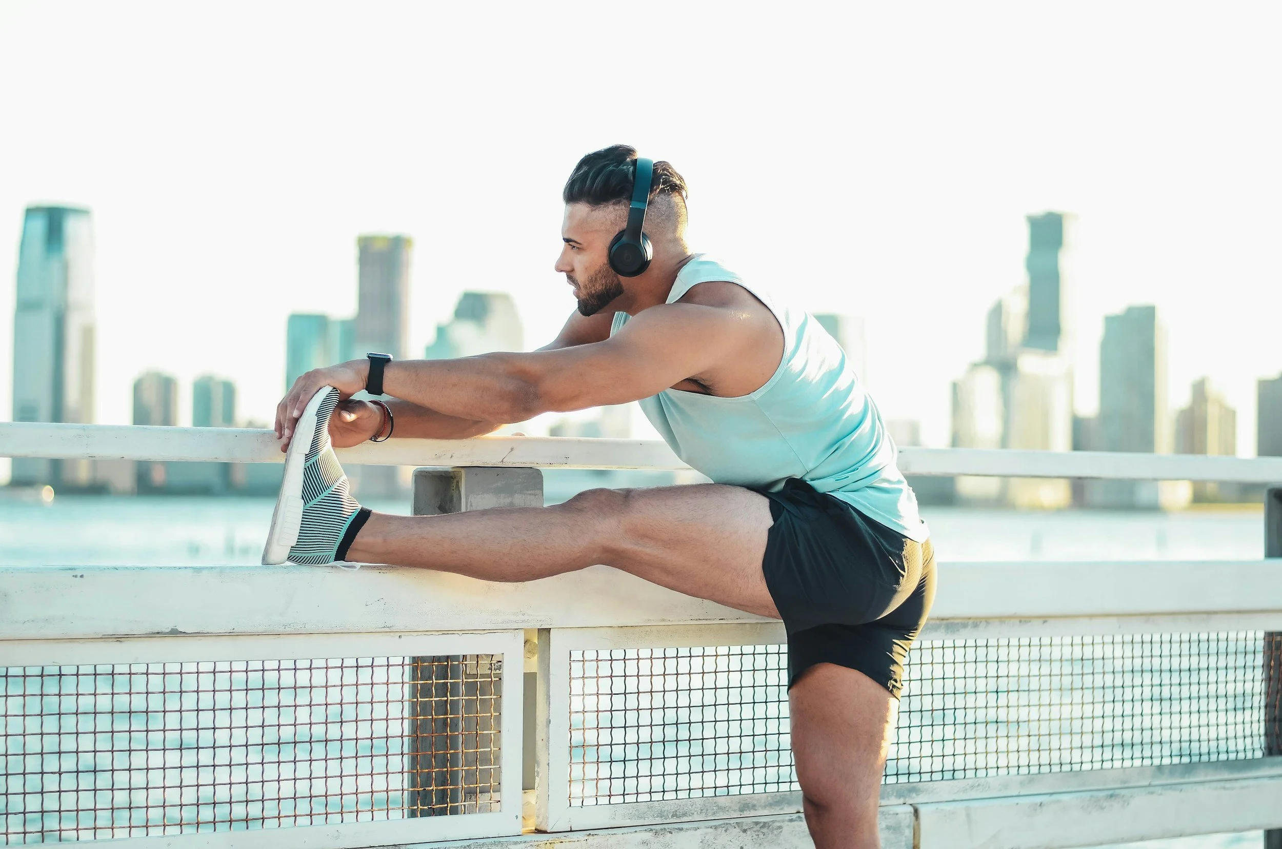 a man wearing a light blue tank top, black shorts, and black headphones stretches with great flexibility on the fence by a river with a downtown skyline in the background. He seems to be suffering from overtraining this spring.