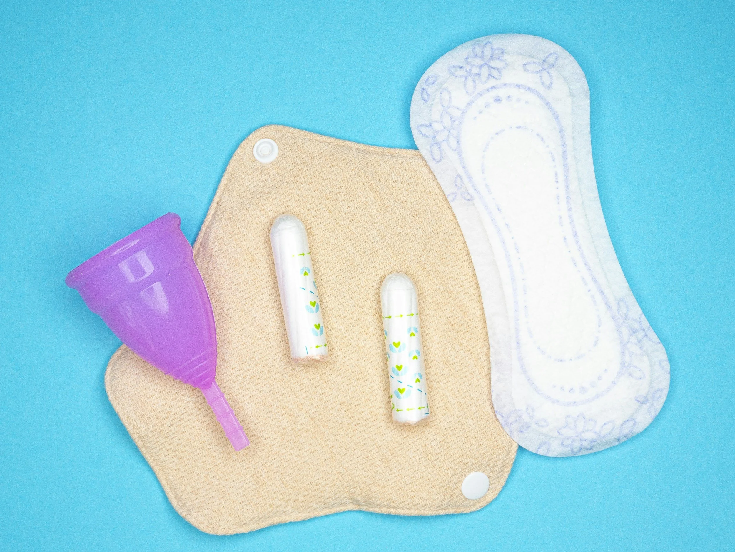 menstrual health products from left to right: menstrual cup, tampons, liner all laying on top of a peach coloured reusable pad.
