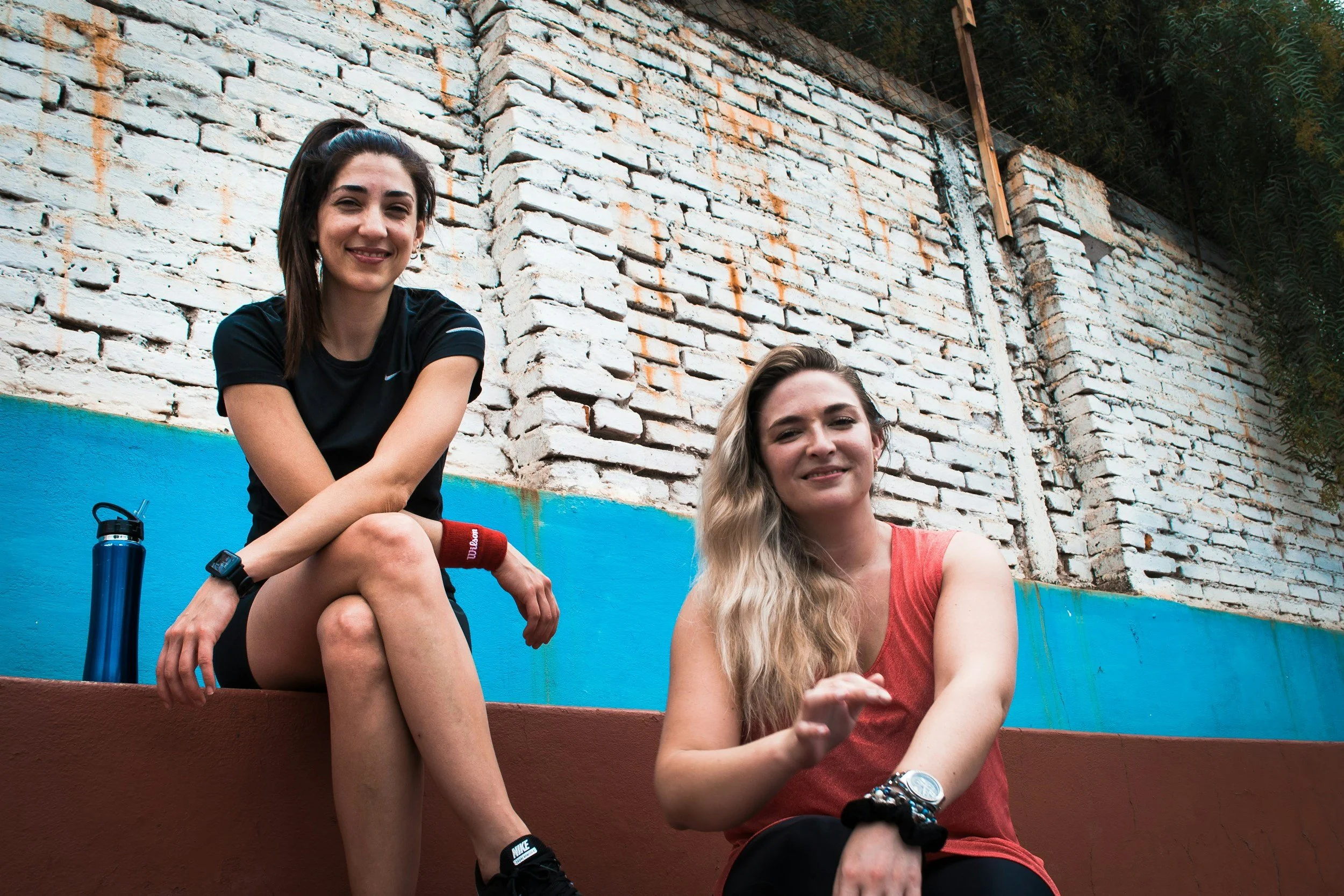 two young women sit in front of a white brick wall with a blue base. They wear sportswear and feel confident about their pelvic health in consideration of their sports since they booked Pelvic Health for Teens with Chantal.