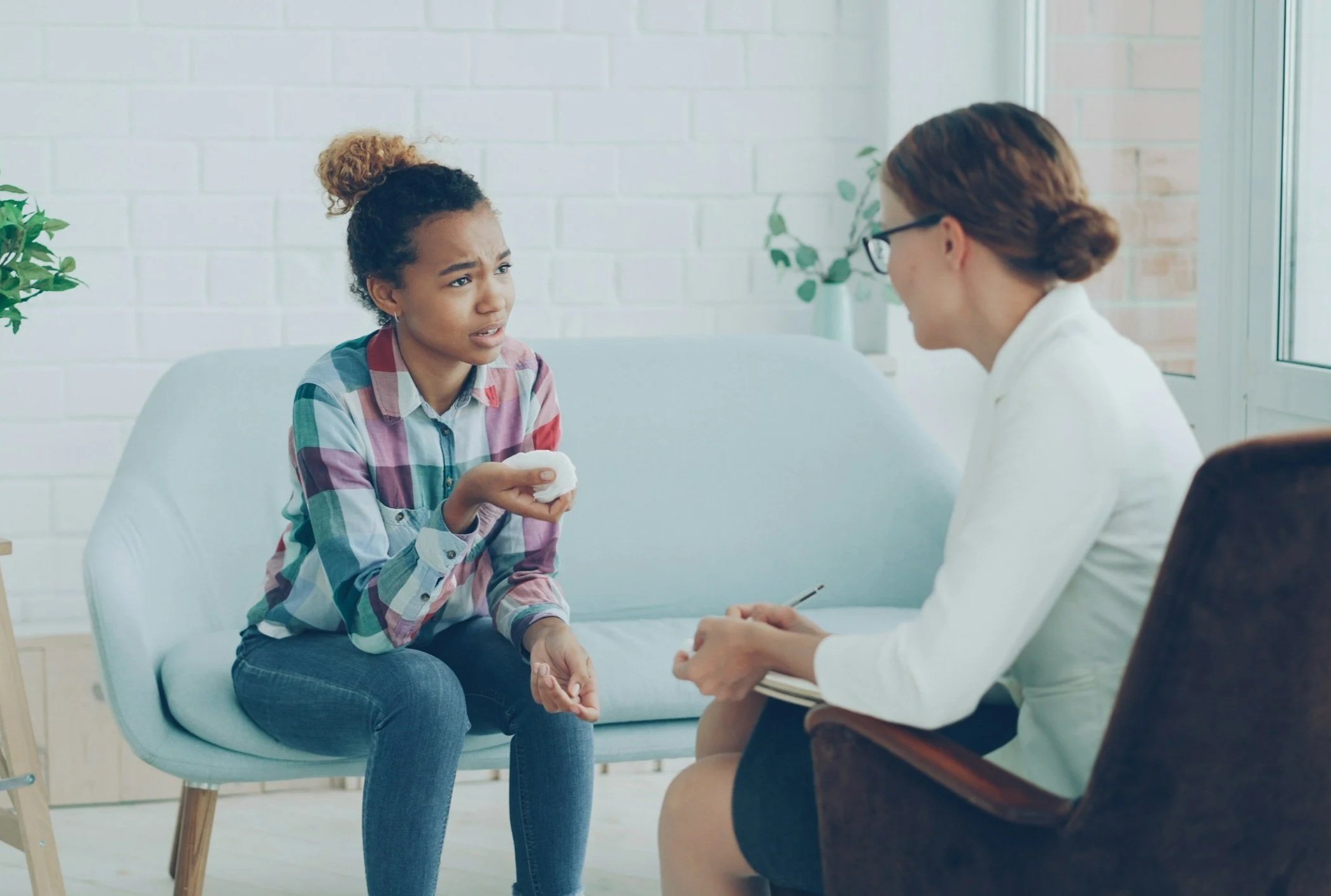 a teen wearing plaid shirt and jeans (left) sits on a pale blue couch and speaks about pelvic health concerns to a professional who is facing away from the viewer (right).