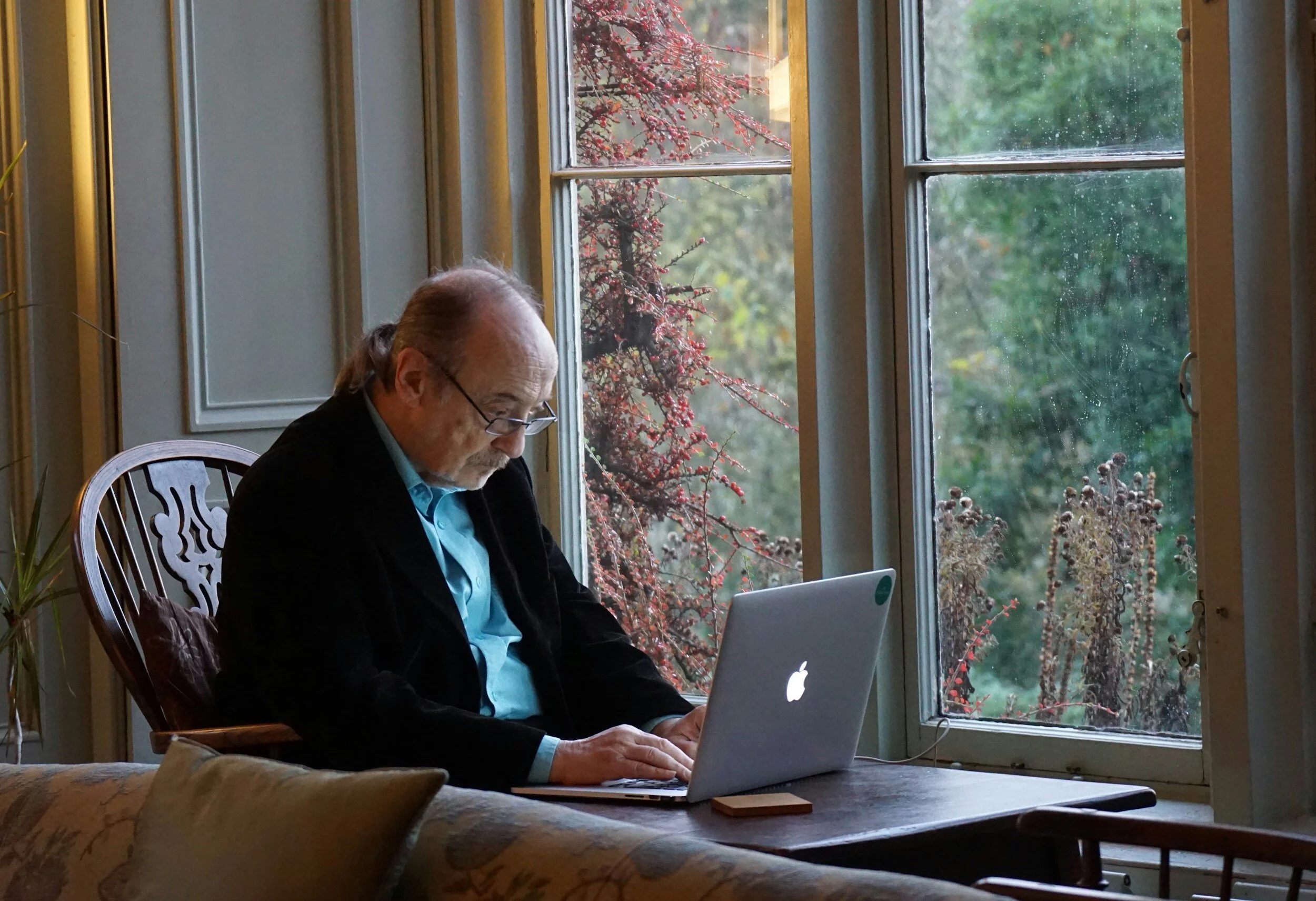 A male enior citizen sitting in from of the window at home using his computer.