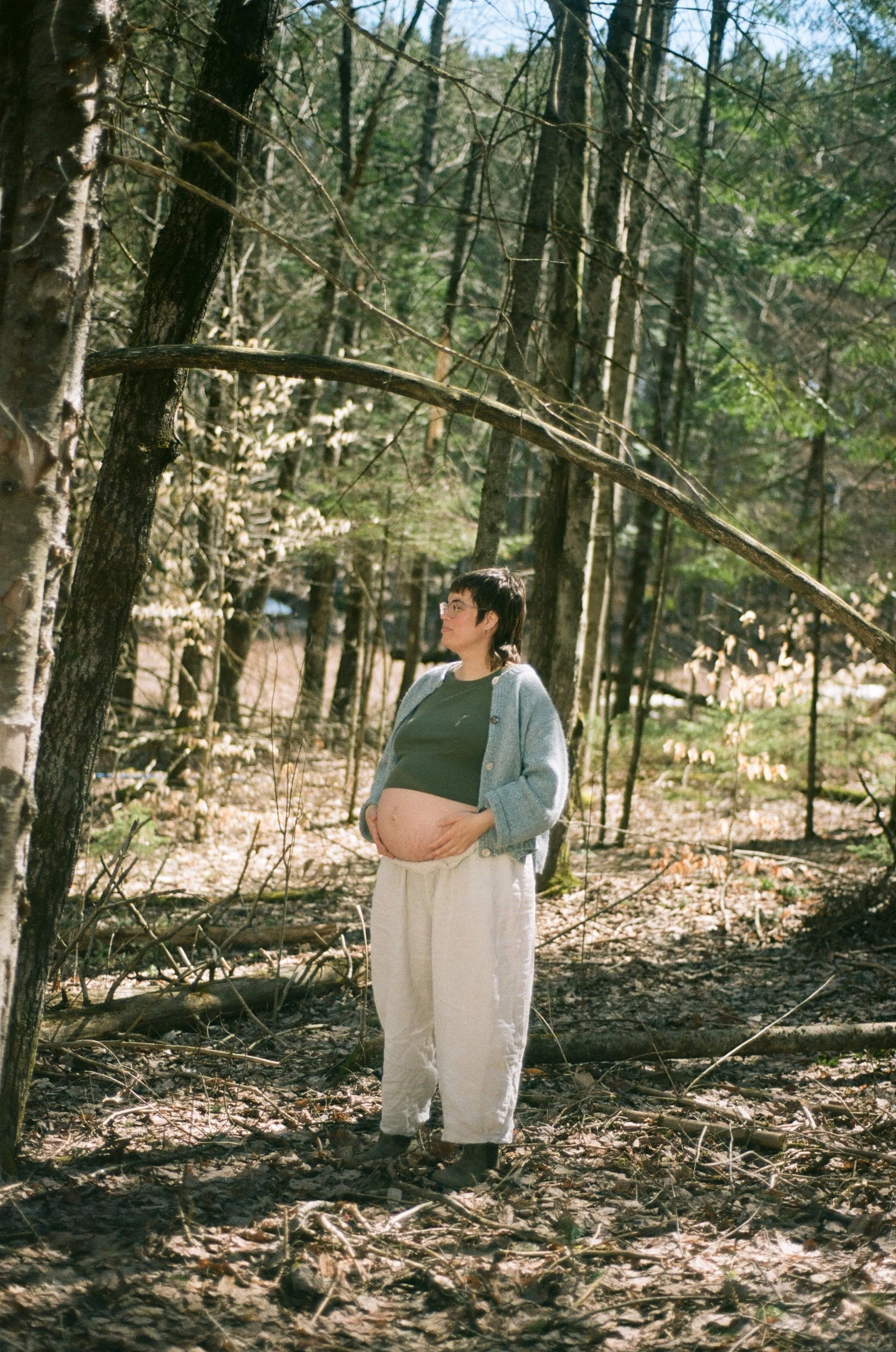 A pregnant person standing in a wooded area during daytime, wearing glasses, a green top, a blue sweater, and beige pants, with their hands resting on her belly and looking to the side.