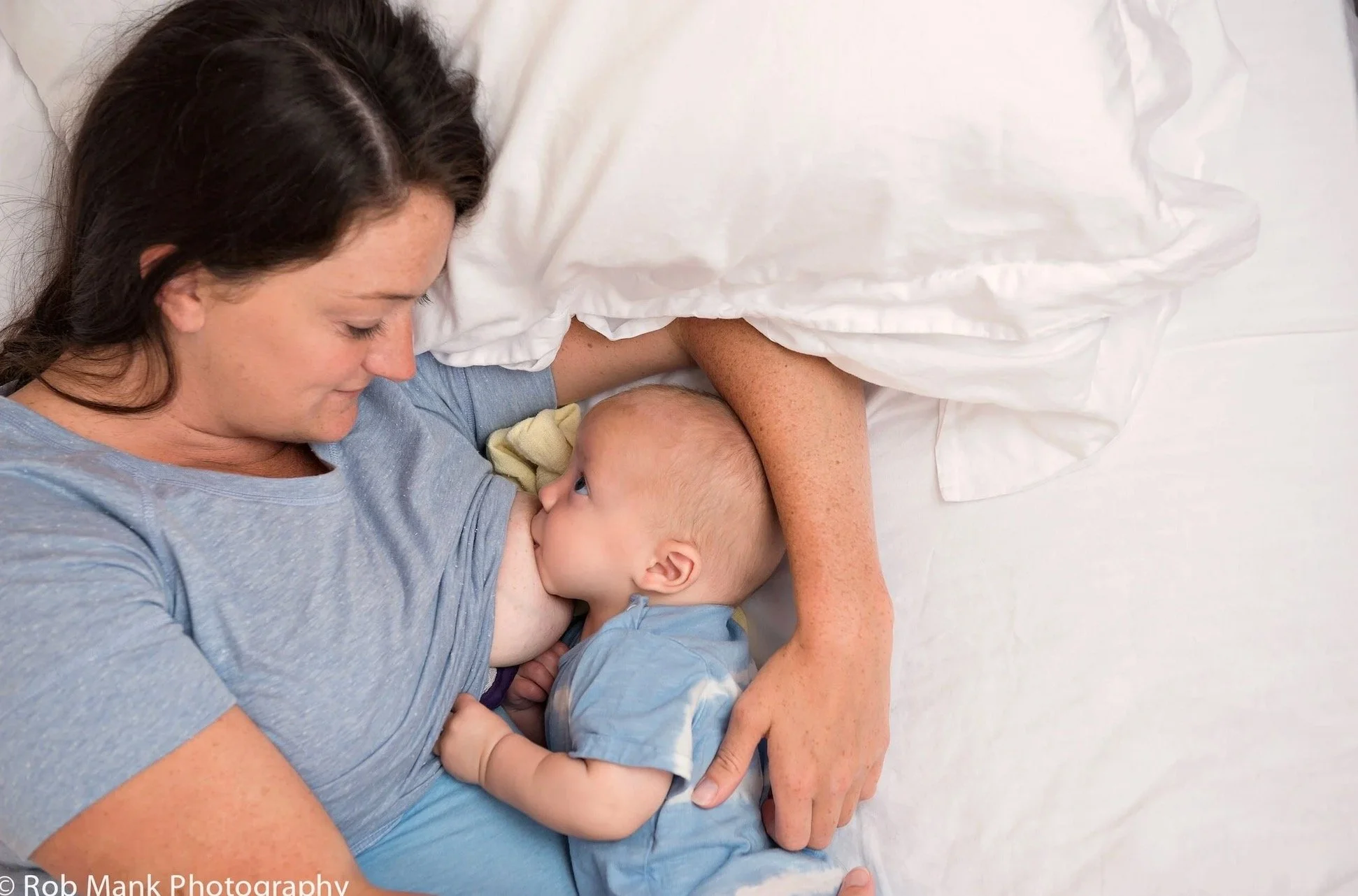 a white person with long hair lying on their side on a white sheet burning a baby who is looking up at them