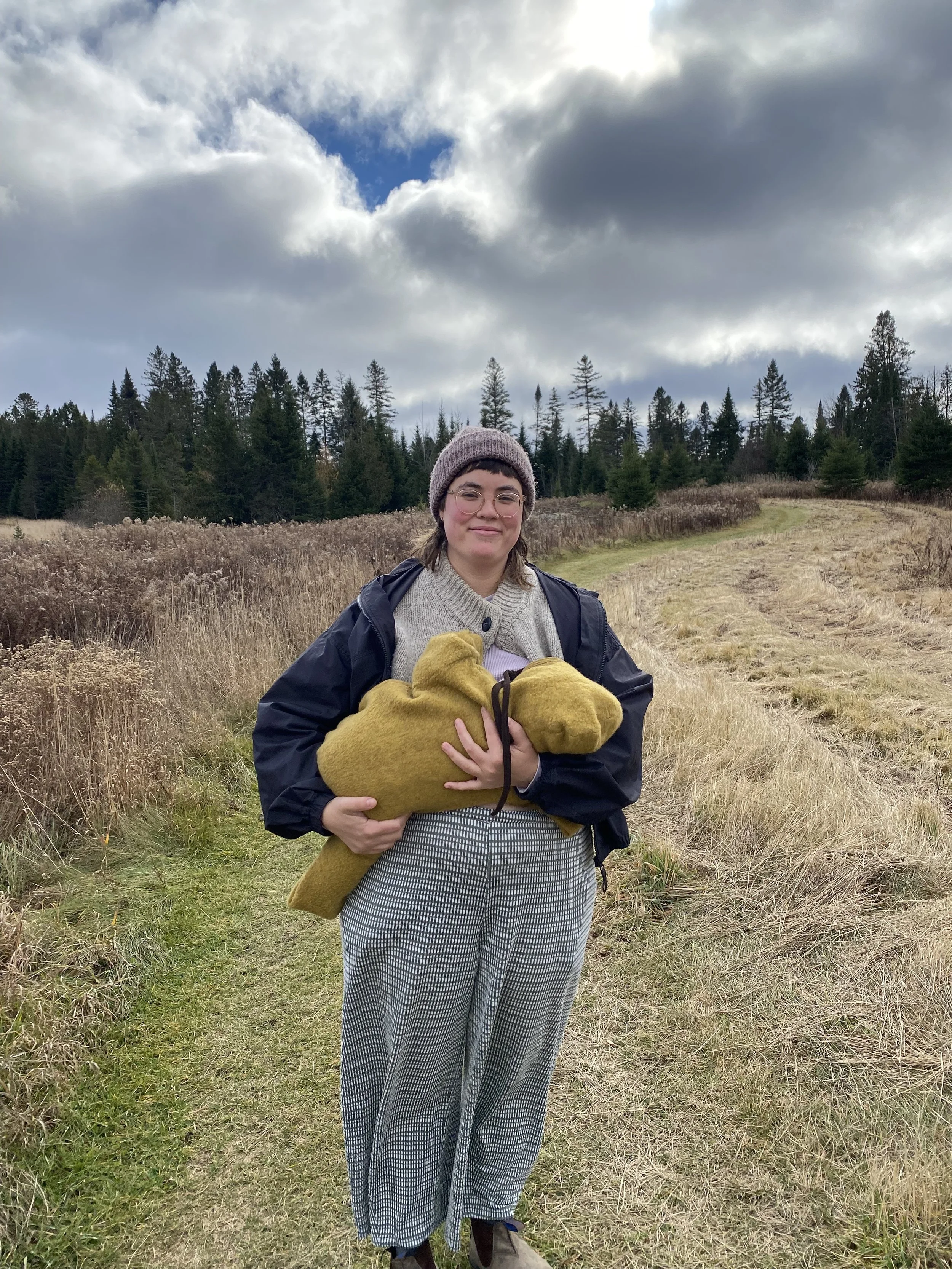 A person standing in a field wearing a hat and jacket and nursing a baby wearing a mustard yellow bunting suit.