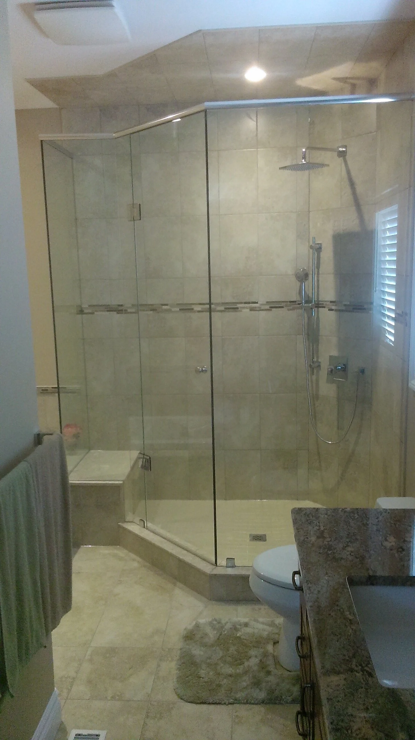 A bathroom with a glass-enclosed shower, a toilet, a granite countertop with a sink, a small rug, and a towel hanging on a rack, with a window with shutters.