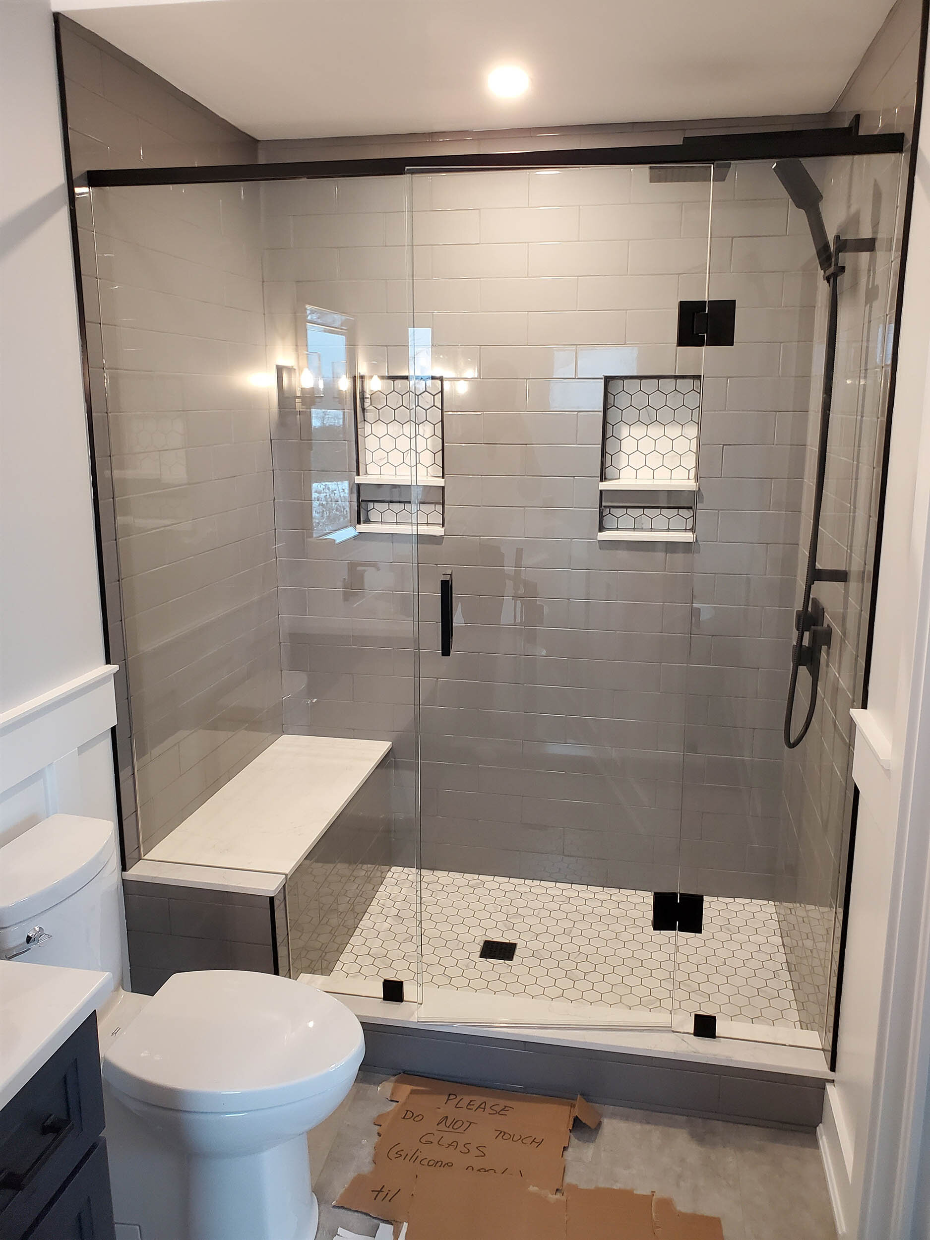 Modern bathroom with a glass-enclosed shower featuring gray subway tiles, two built-in shelves with tile backs, and a black showerhead and fixtures. A white toilet and a small white cabinet are visible, along with a cardboard sign on the floor that r