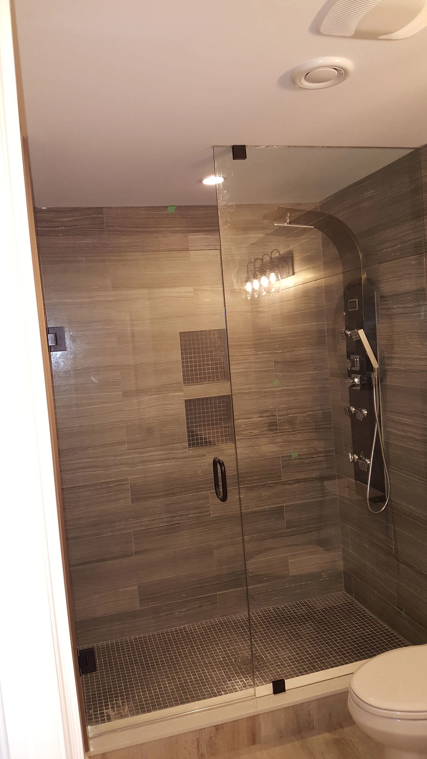 A modern bathroom shower area with glass enclosure, brown tiled walls, black accents, and a built-in shelf with small square tiles.