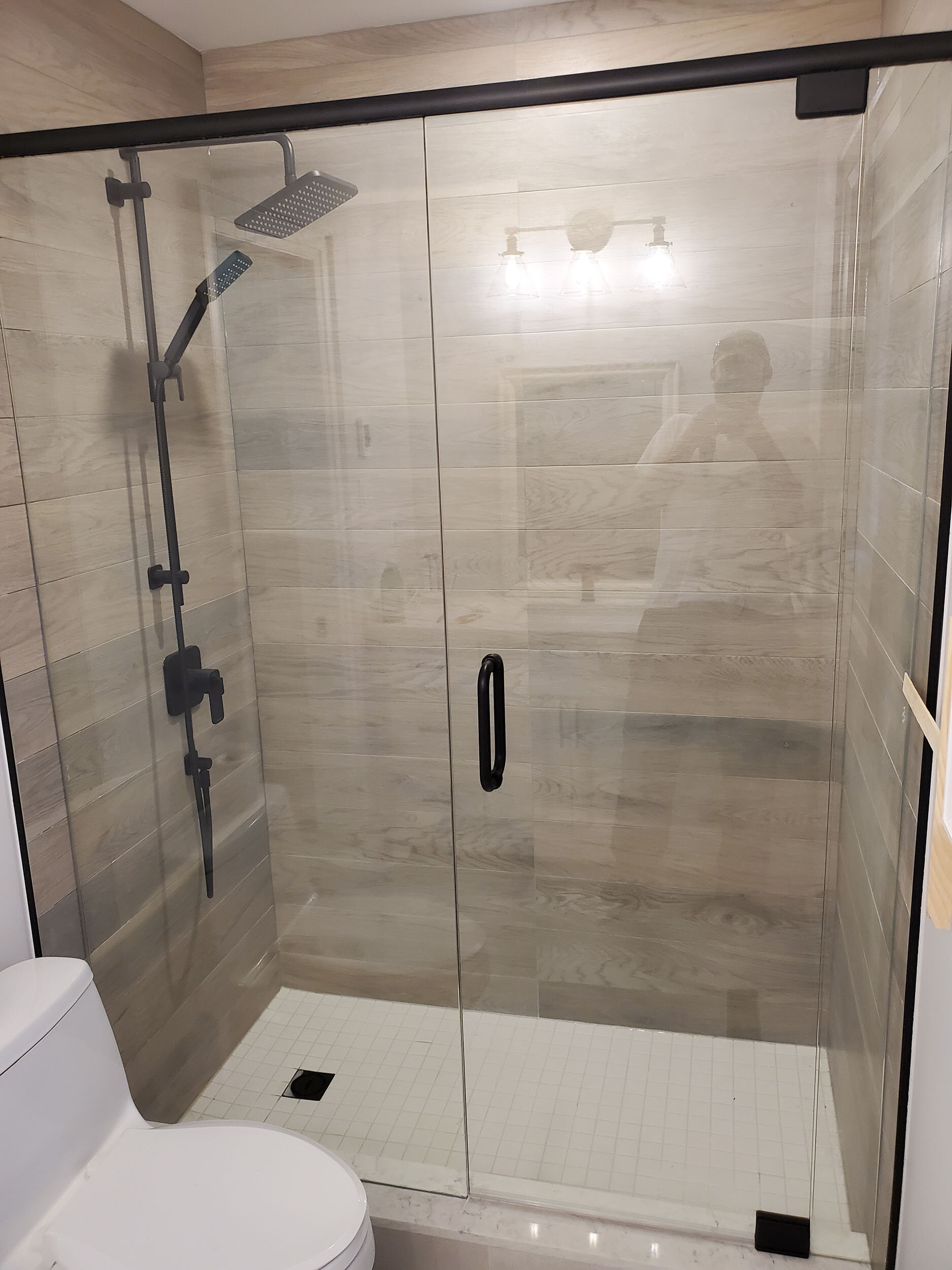 A modern glass-enclosed shower with beige tiled walls, a black handle on the door, a silver overhead rain shower, and a smaller handheld shower attached to a vertical bar. A reflection of a person taking the photo is visible in the glass, along with 