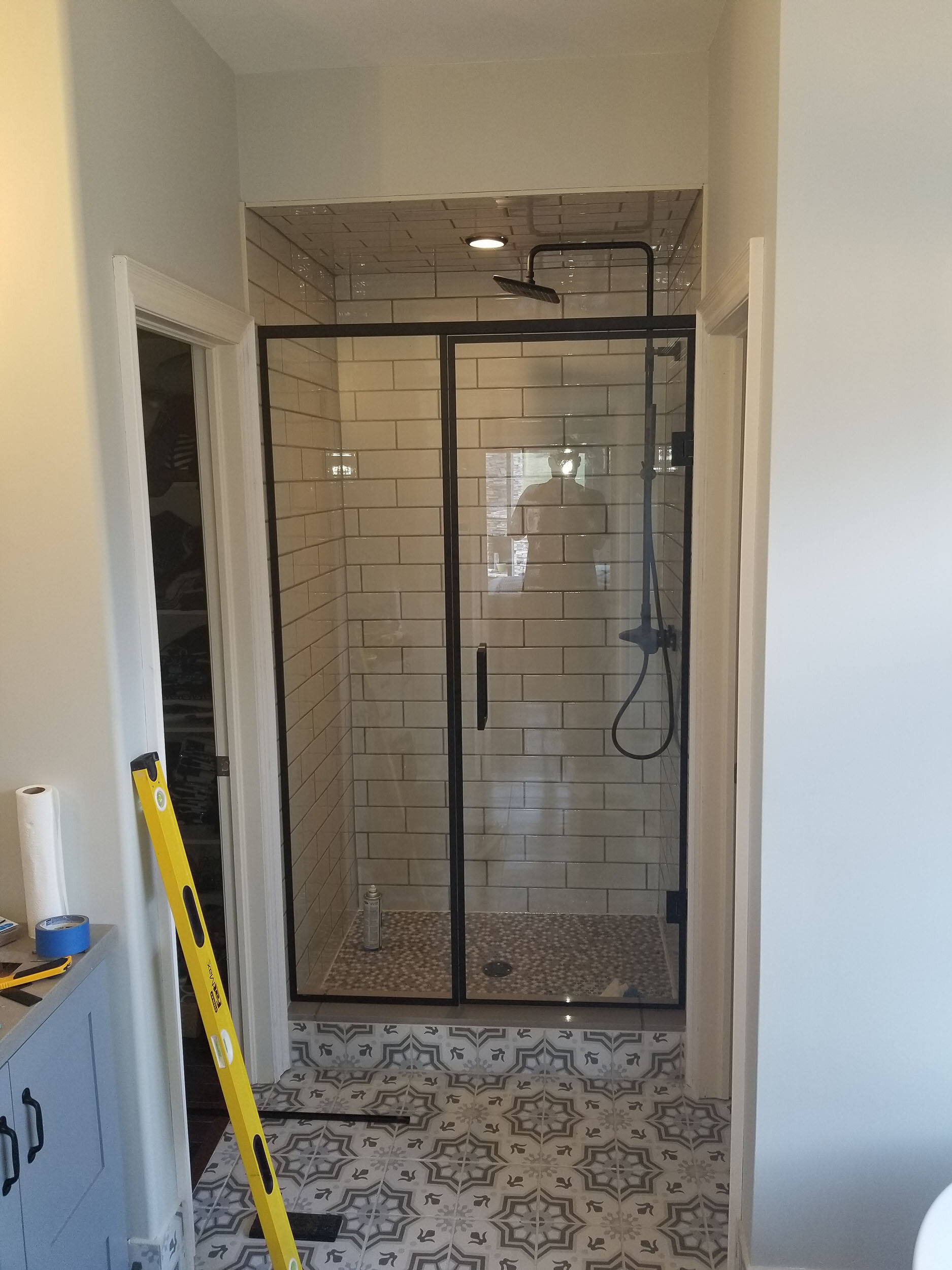 A modern walk-in shower with glass door and black metal framing, white subway tiles on the walls, a rainfall showerhead, and decorative patterned floor tiles. Reflection of a person taking the photo is visible in the glass.