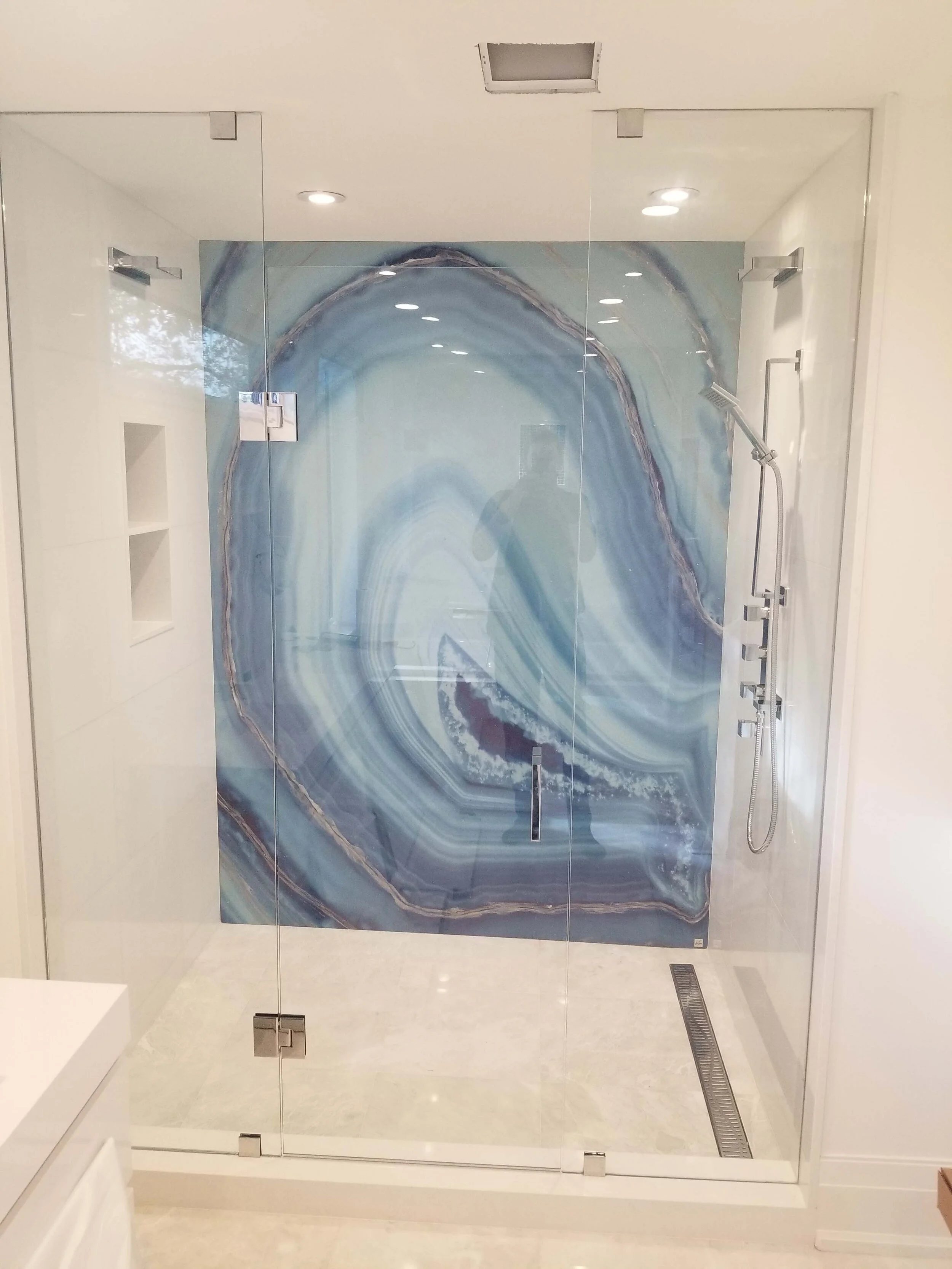 Modern shower with a large blue and white marble-patterned accent wall, glass enclosure, corner shower head, built-in shelves, and ceiling lights.