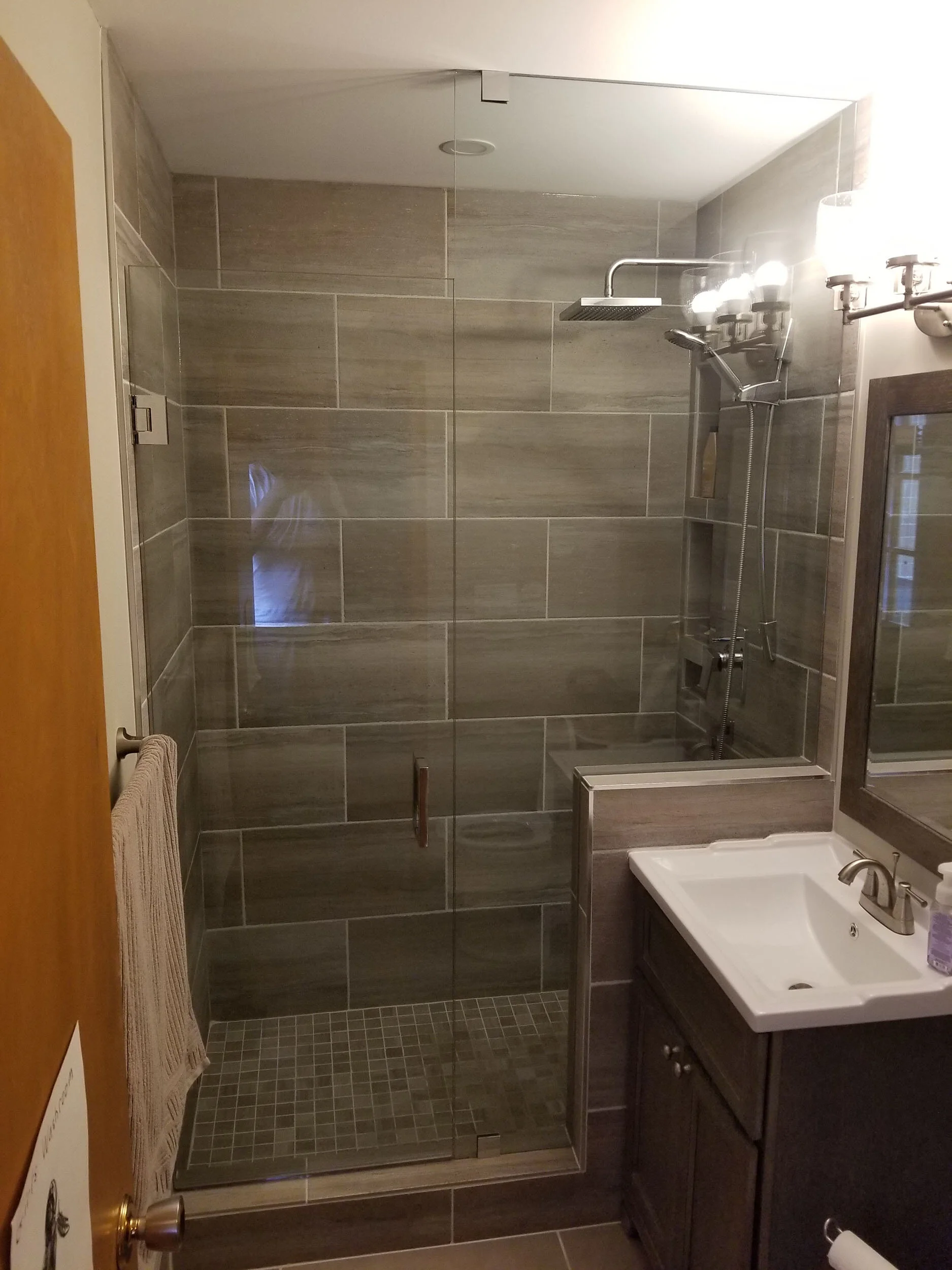 Modern bathroom with glass-enclosed shower, grey tiled walls and floor, a white sink with dark cabinet, and a mirror with lights.