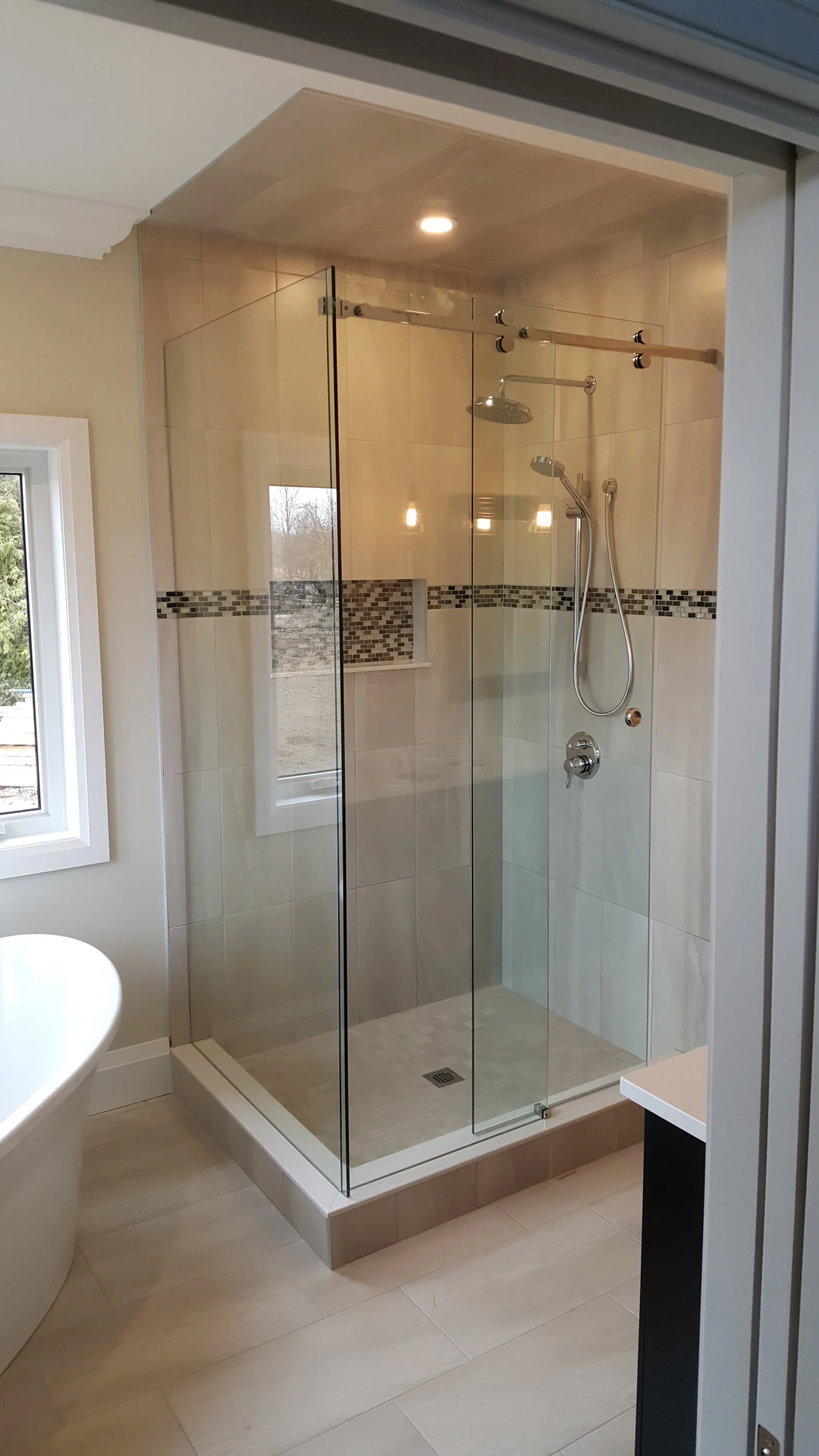 Modern bathroom with a glass-enclosed shower featuring beige tiles and a decorative mosaic strip, a window, and a light overhead.