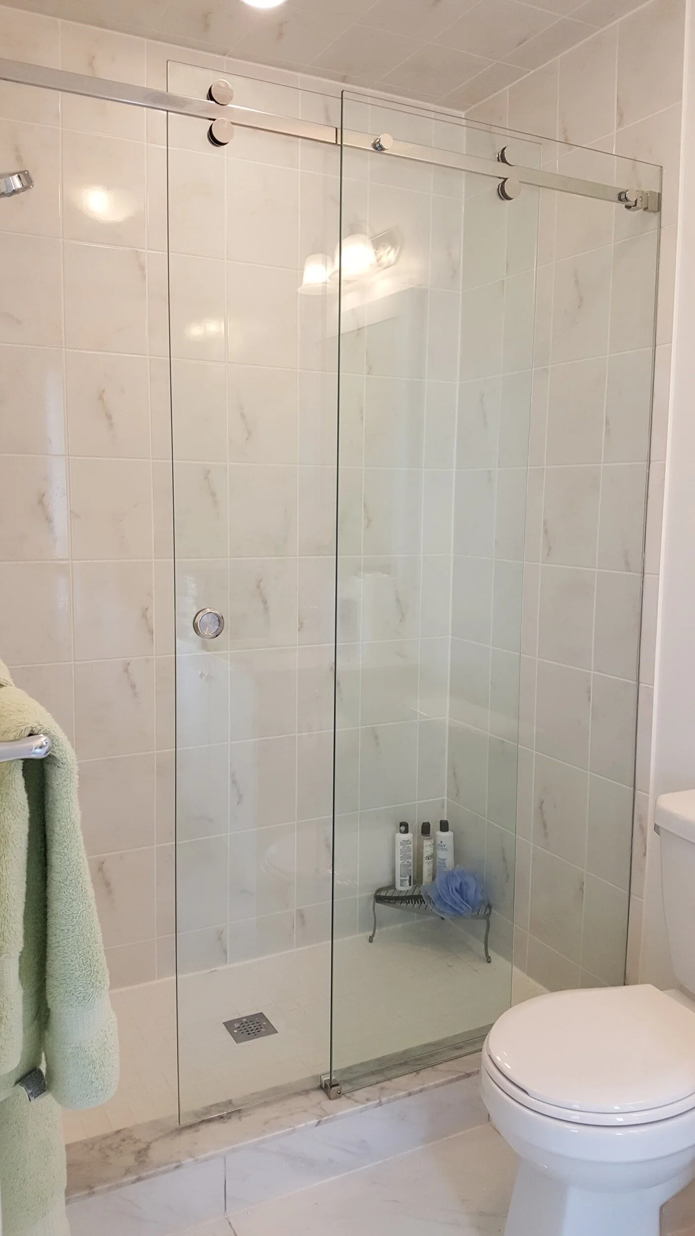 Bathroom with a glass shower enclosure, marble tiles, a towel on a rack, and toiletries on a small stand inside the shower.