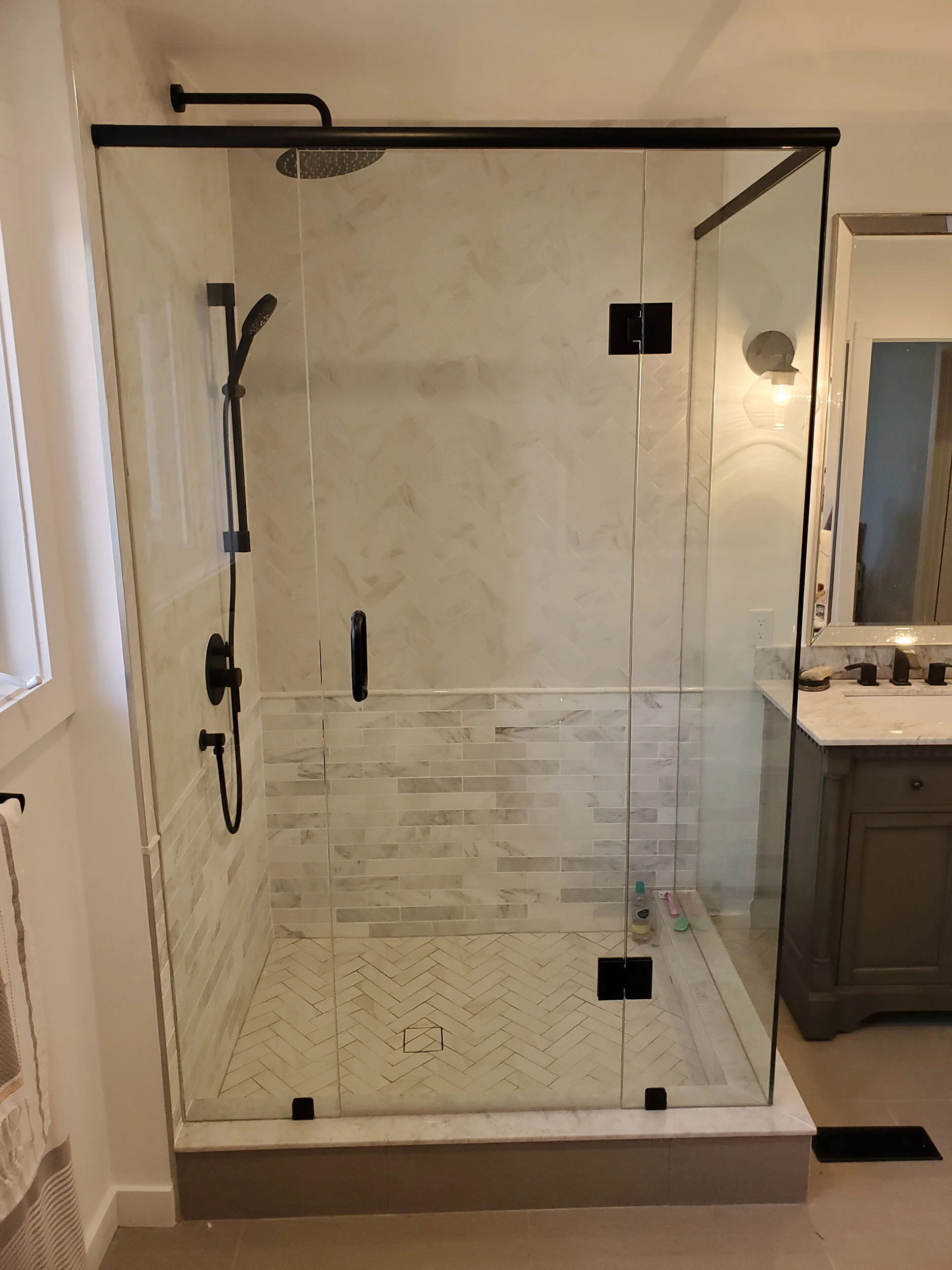 A modern walk-in shower with glass door and black hardware, tiled walls, and a drain on the floor, adjacent to a bathroom vanity with a marble countertop and mirror.