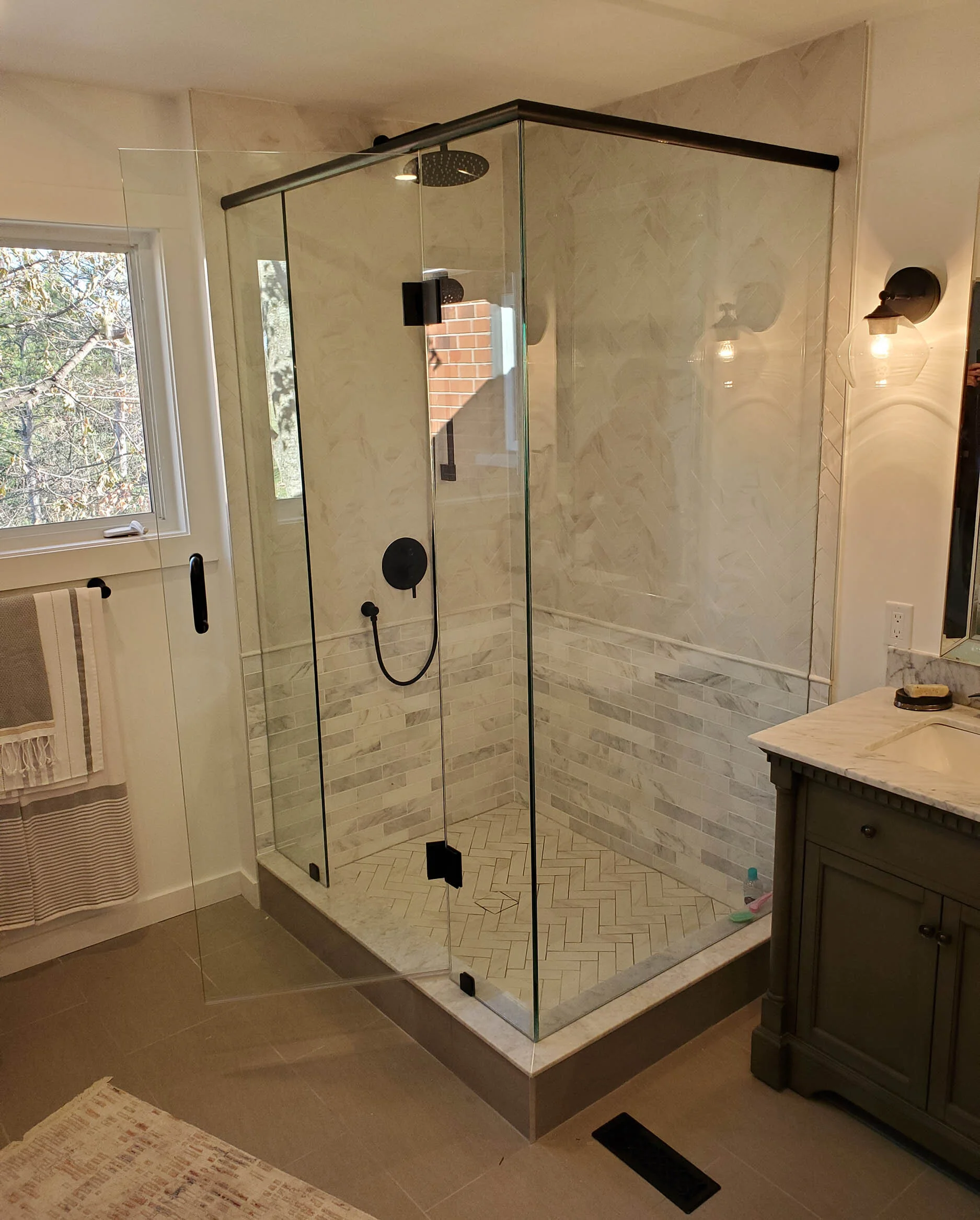 A modern glass shower enclosure with a black rain shower head and a black handheld shower in a bathroom with a window, bathroom vanity, and beige tile flooring.