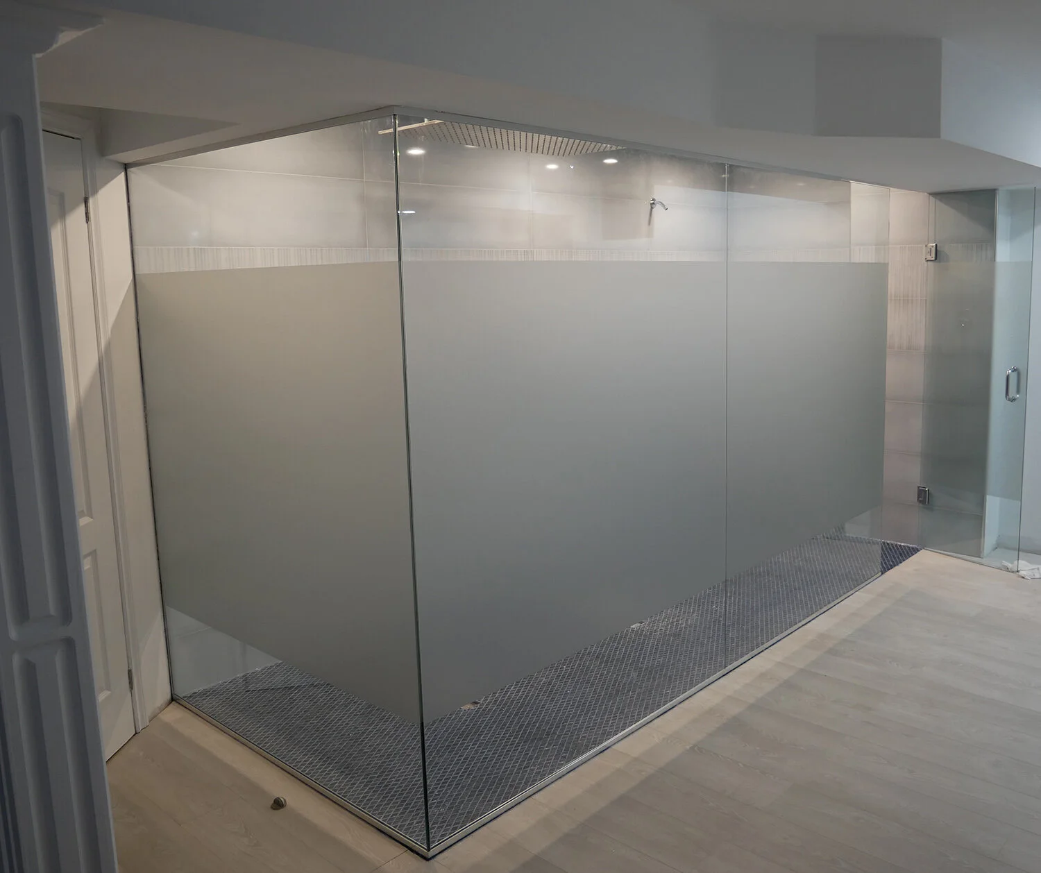 Frosted glass enclosure in a modern shower room with a tiled wall, ceiling lights, and a drain on the floor.