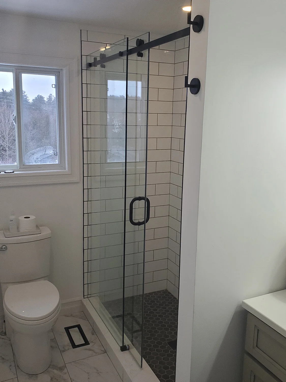 A modern bathroom with a glass shower enclosure, white tiled walls, and a black shower frame. There is a window with a view of a snowy outdoor scene, a toilet, and a marble floor.