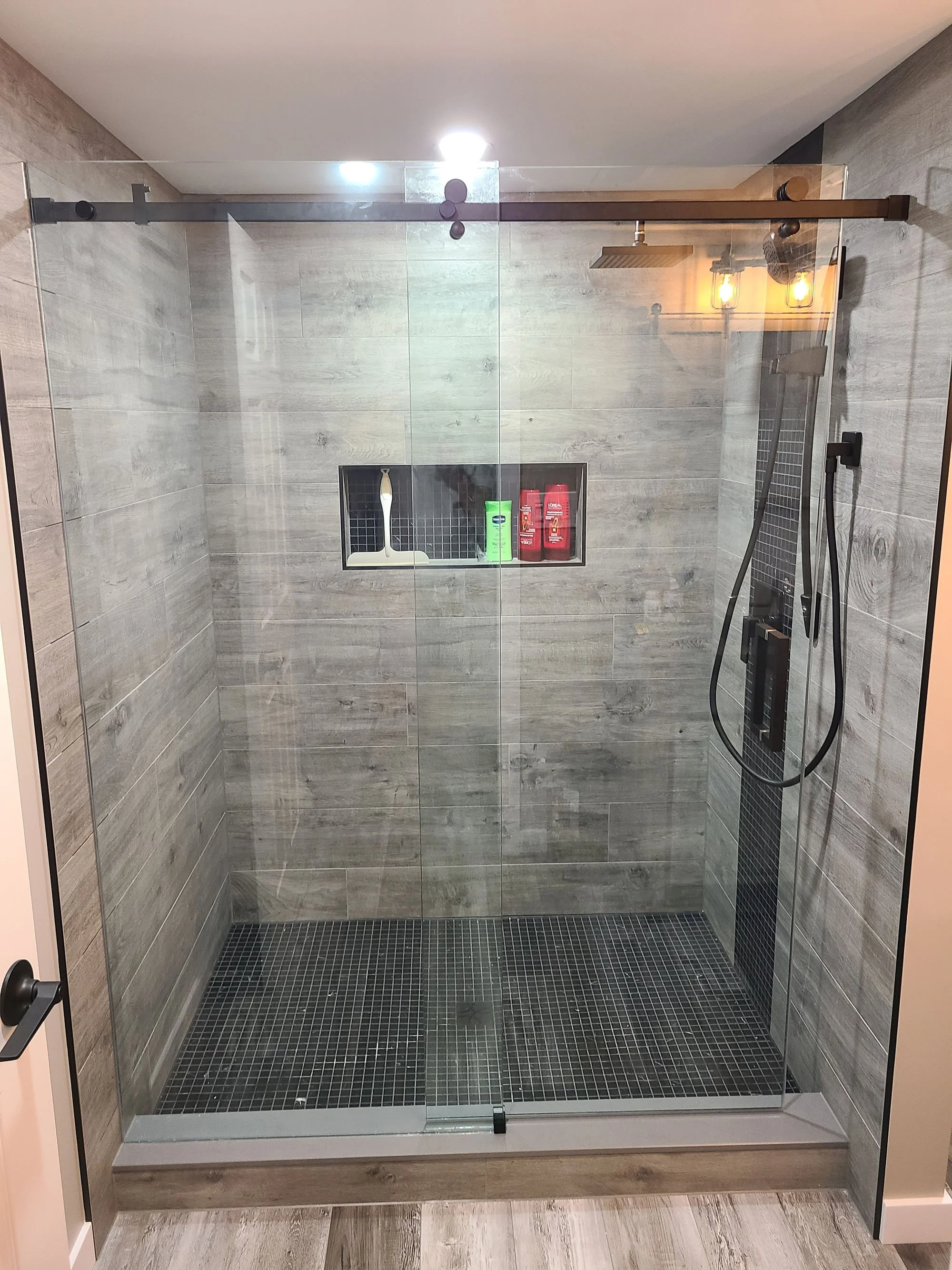 Modern walk-in shower with glass doors, wood-look tiles on walls and floor, black mosaic tiles on floor, recessed niche with toiletries, overhead rain shower, and black hand-held shower head.