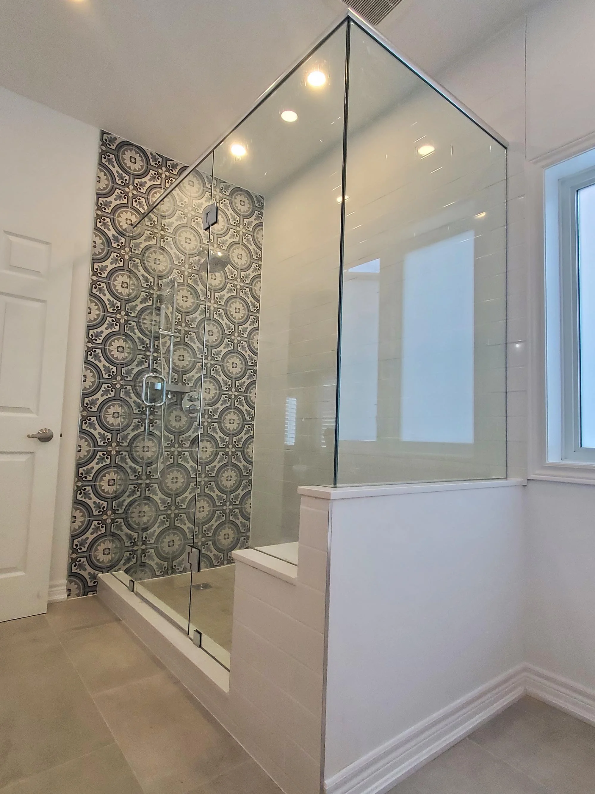 Bathroom shower with clear glass enclosure, decorative patterned tiles on the back wall, beige floor tiles, and a window letting in natural light.
