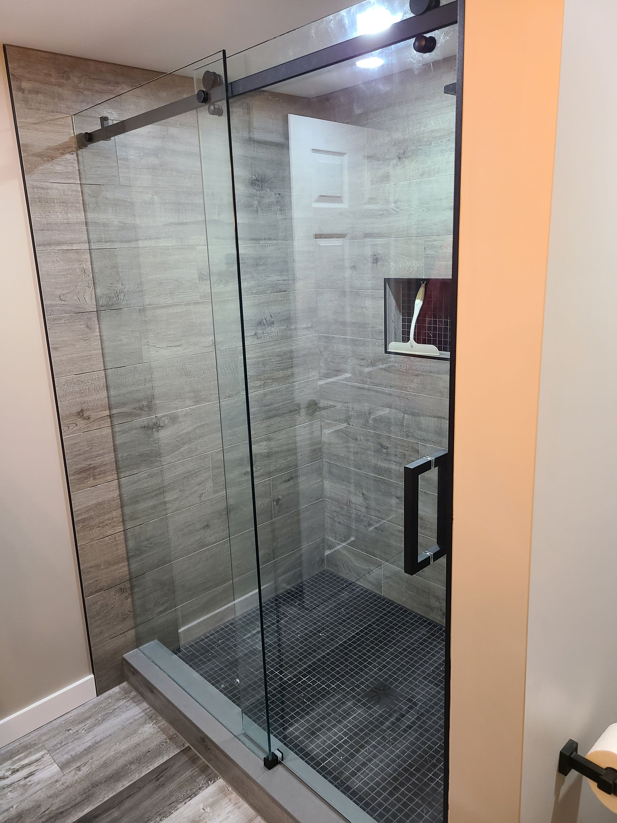 A modern glass shower enclosure with black hardware, wood-look tile walls, black mosaic tile floor, a built-in niche, and a brushed nickel threshold at the entrance.