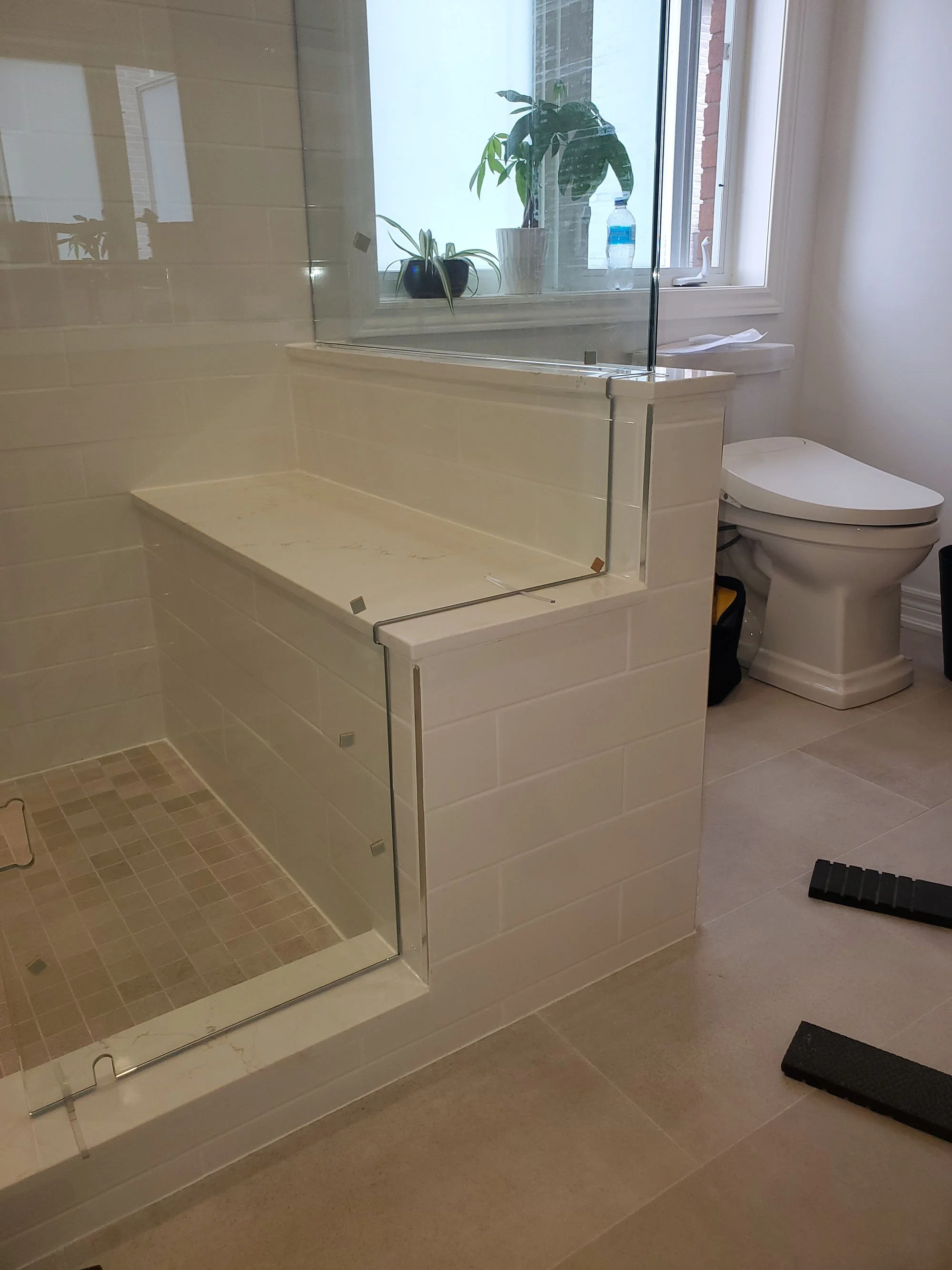Bathroom with walk-in shower enclosed by glass with a marble bench, and a toilet in front of a window with potted plants.
