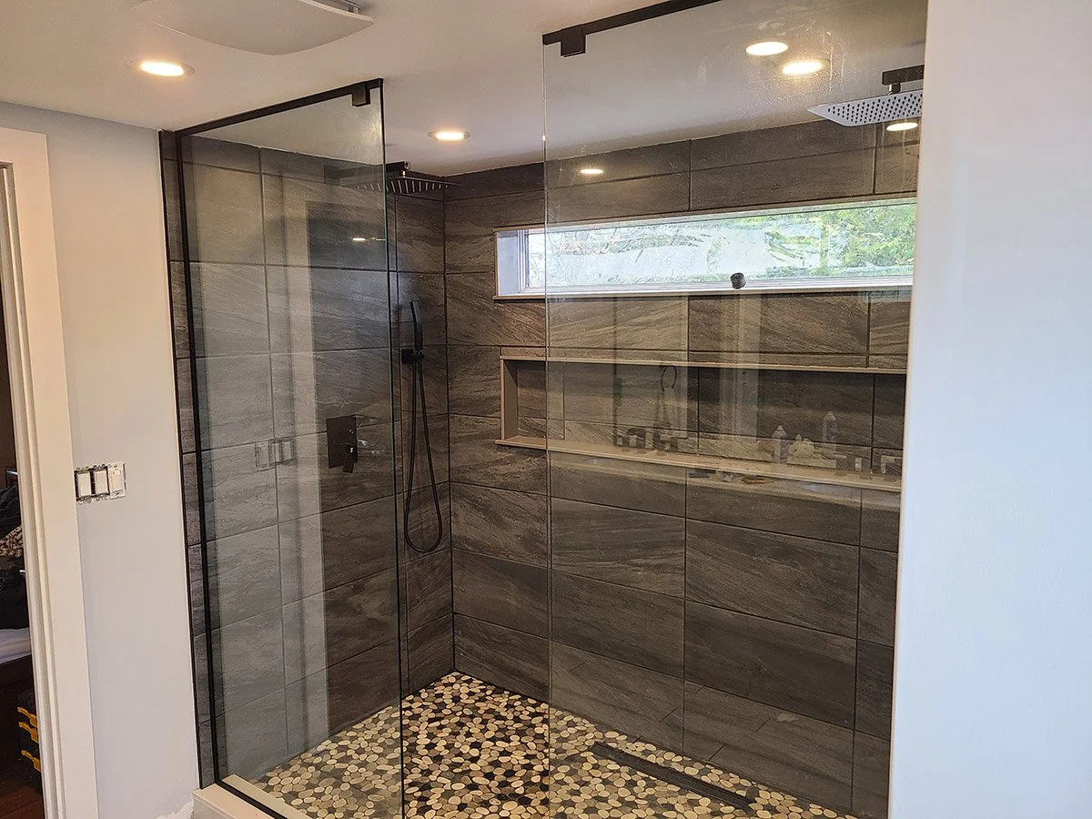 Modern walk-in shower with glass panels, gray stone tiles on the walls and pebble tiles on the floor, and a slim horizontal window for natural light.