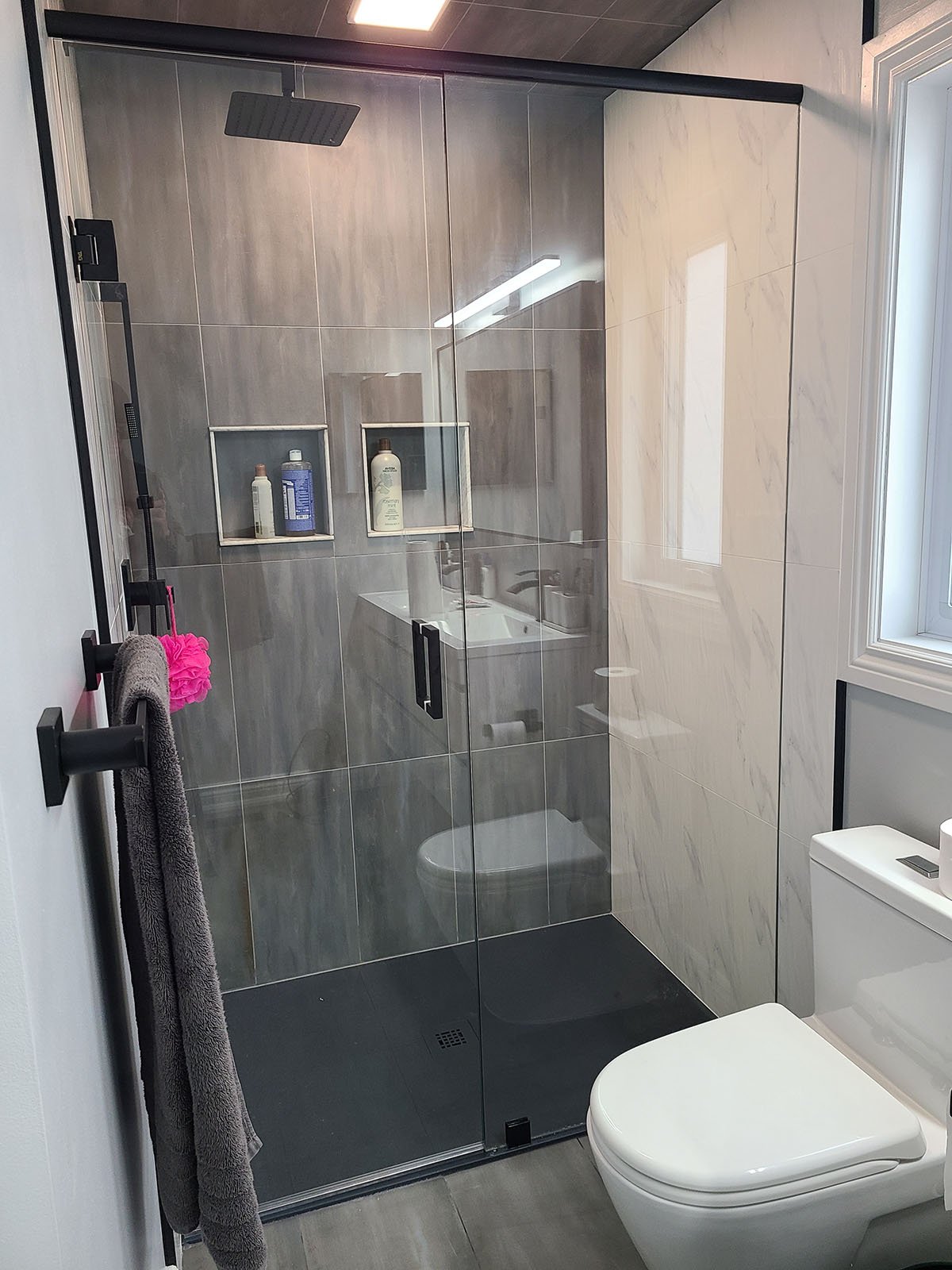 Modern bathroom with a glass shower enclosure, grey and beige tiles, a wall-mounted towel rack with a grey towel and pink scrubber, a white toilet, and a window allowing natural light.